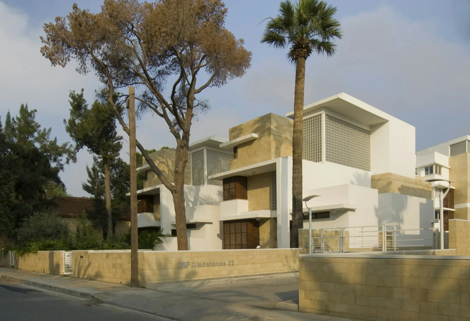 Modern residential building with geometric white and golden-tan facades, featuring a palm tree and mature olive tree in the foreground, designed by Theo.David Architects.