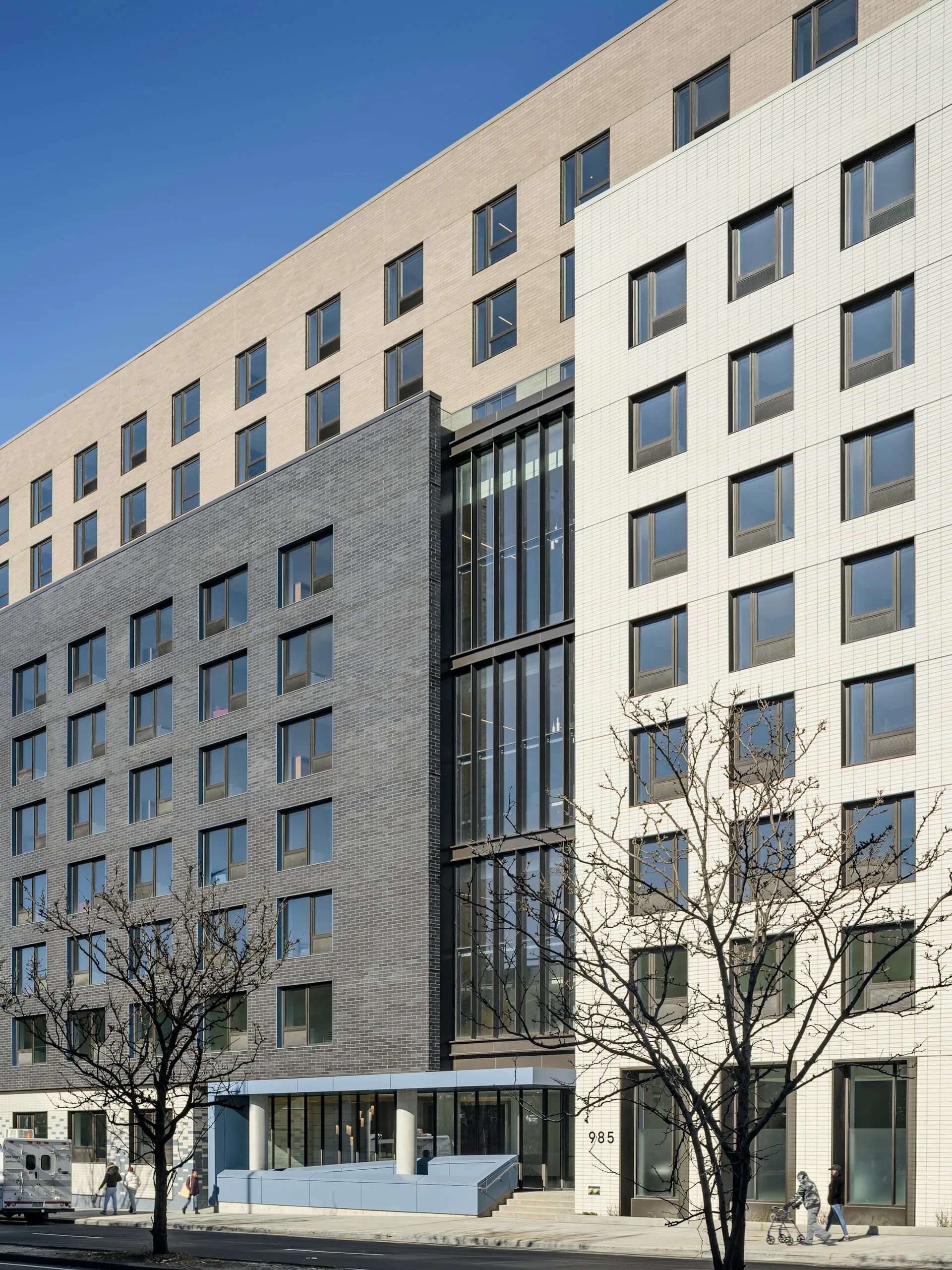 Modern multi-story residential building with mixed materials—light-colored brick upper floors and darker brick lower section—featuring regularly spaced windows and a dramatic vertical glass entry feature, designed by Think! Architecture + Design.