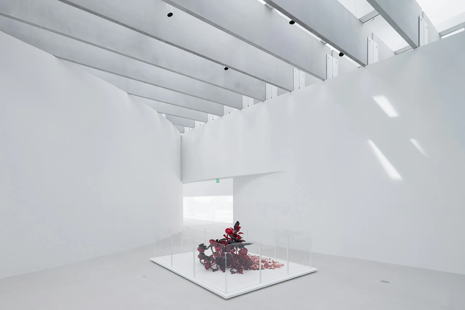 Minimalist white gallery space with exposed beam ceiling and skylight, featuring a small sculptural installation with red elements on a central platform by Thomas Phifer and Partners.