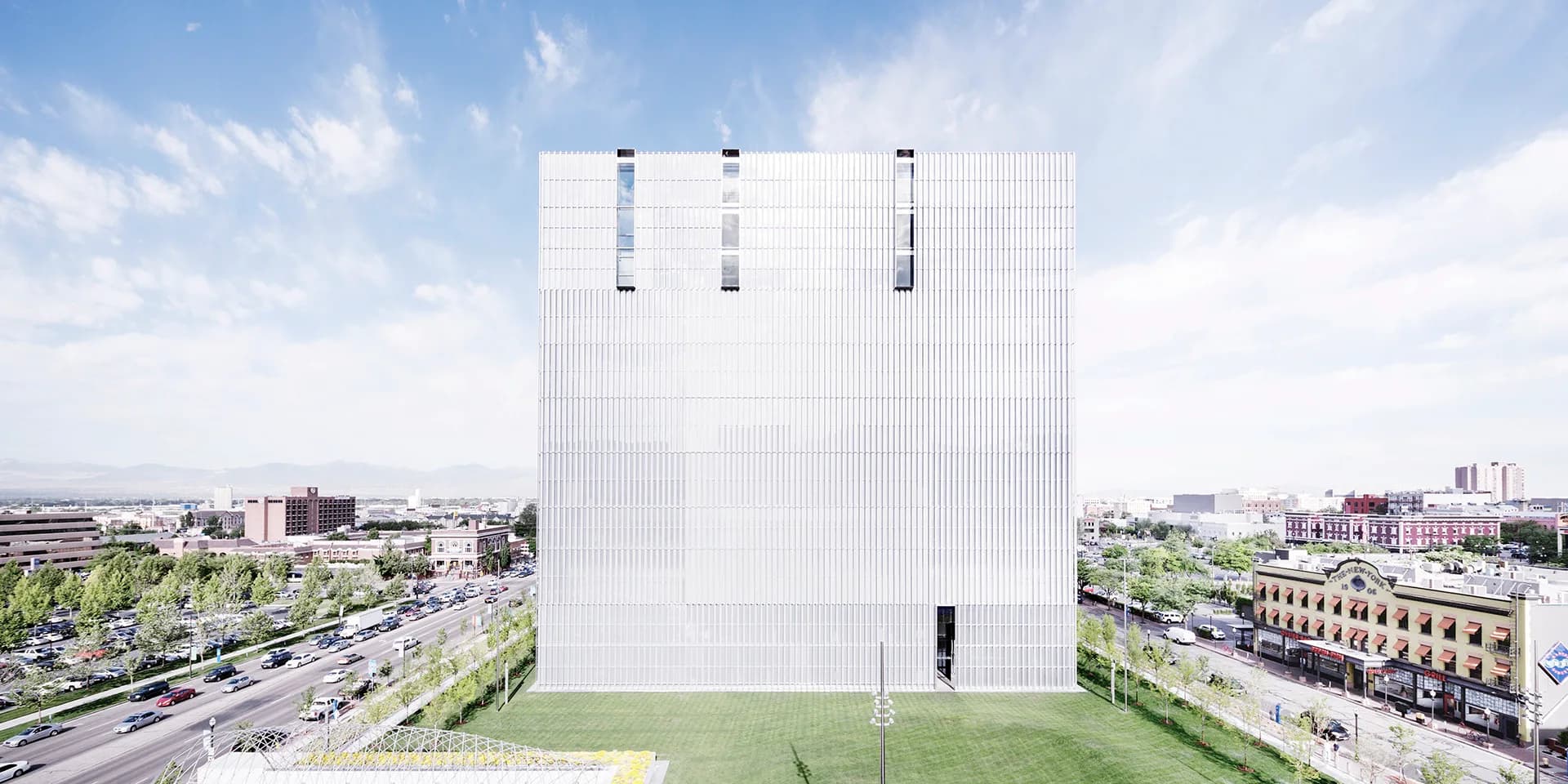 A white modernist rectangular tower with three vertical window slits rises above green landscaping, photographed against a cloudy sky with urban cityscape beyond, by Thomas Phifer and Partners.