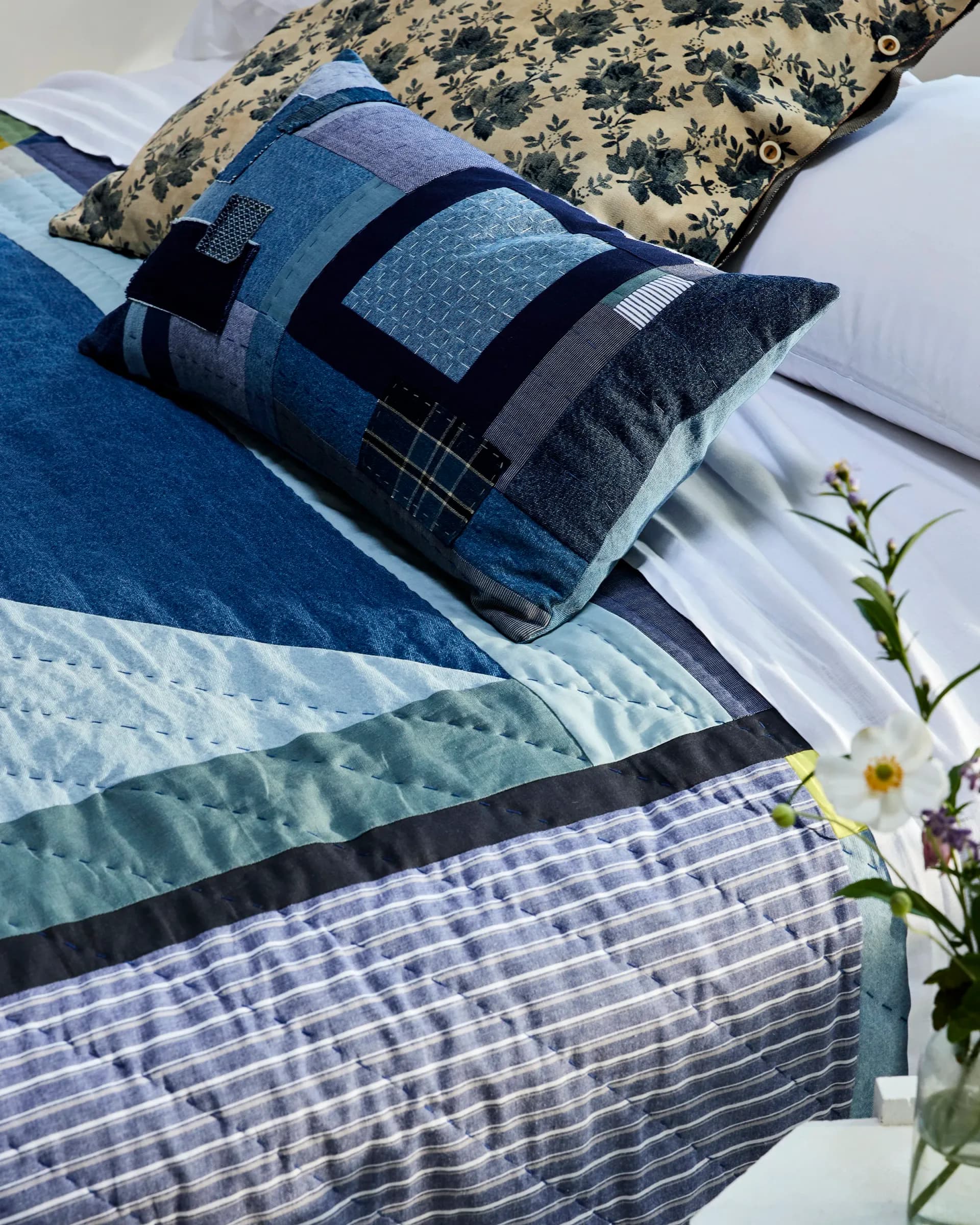 Styled bedroom display featuring layered blue and patterned pillows, quilted bedding, and fresh flowers in a neutral palette by Thompson Street Studio.
