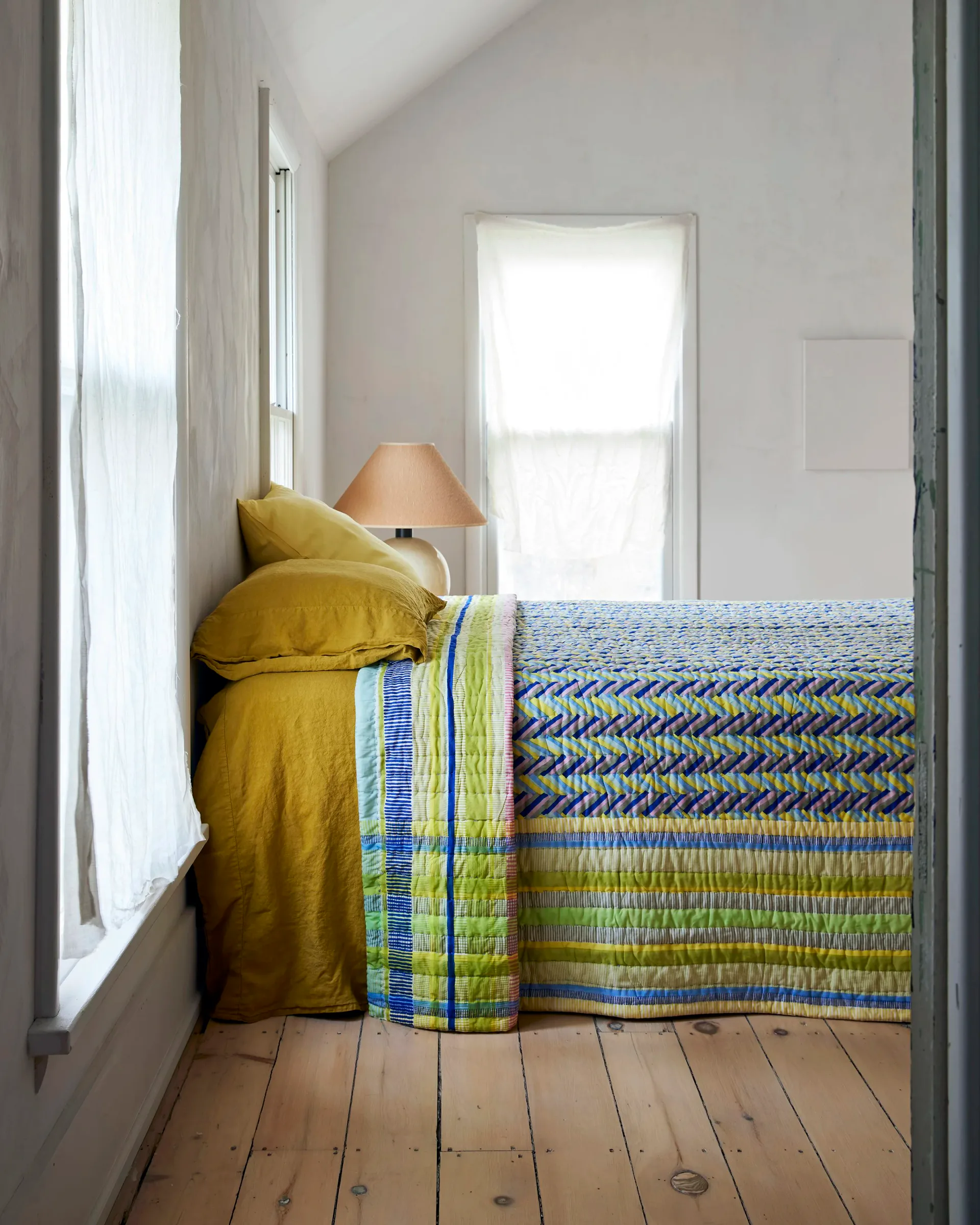 Bright bedroom with mustard-yellow bedding, woven blue and green throw blanket, wooden floors, and abundant natural light from windows by Thompson Street Studio.