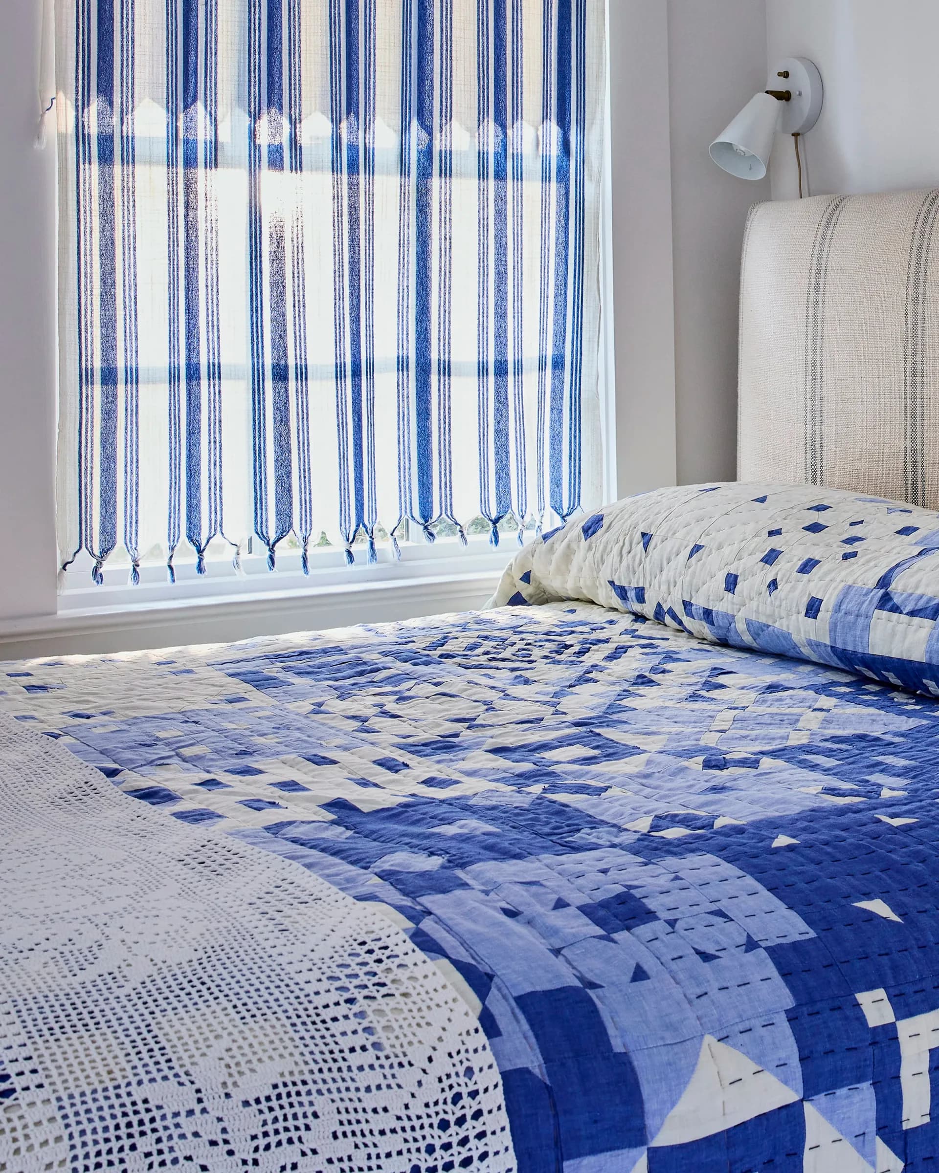 Bedroom interior featuring blue and white patterned bedding, striped vertical blinds, and neutral wall-mounted headboard by Thompson Street Studio.