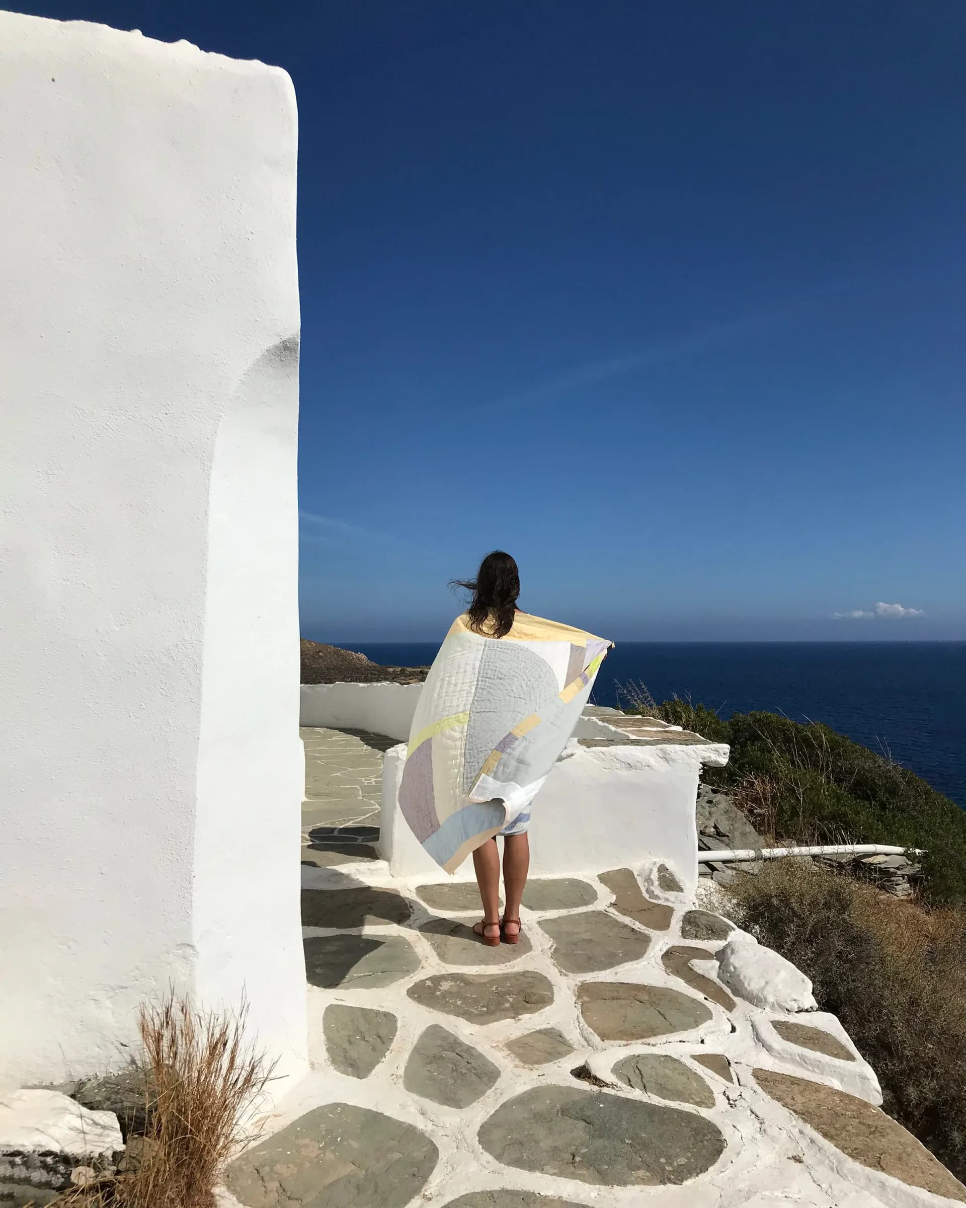Whitewashed Mediterranean terrace with stone flooring and white walls overlooking the sea, featuring minimal furnishings and natural coastal landscape by Thompson Street Studio.