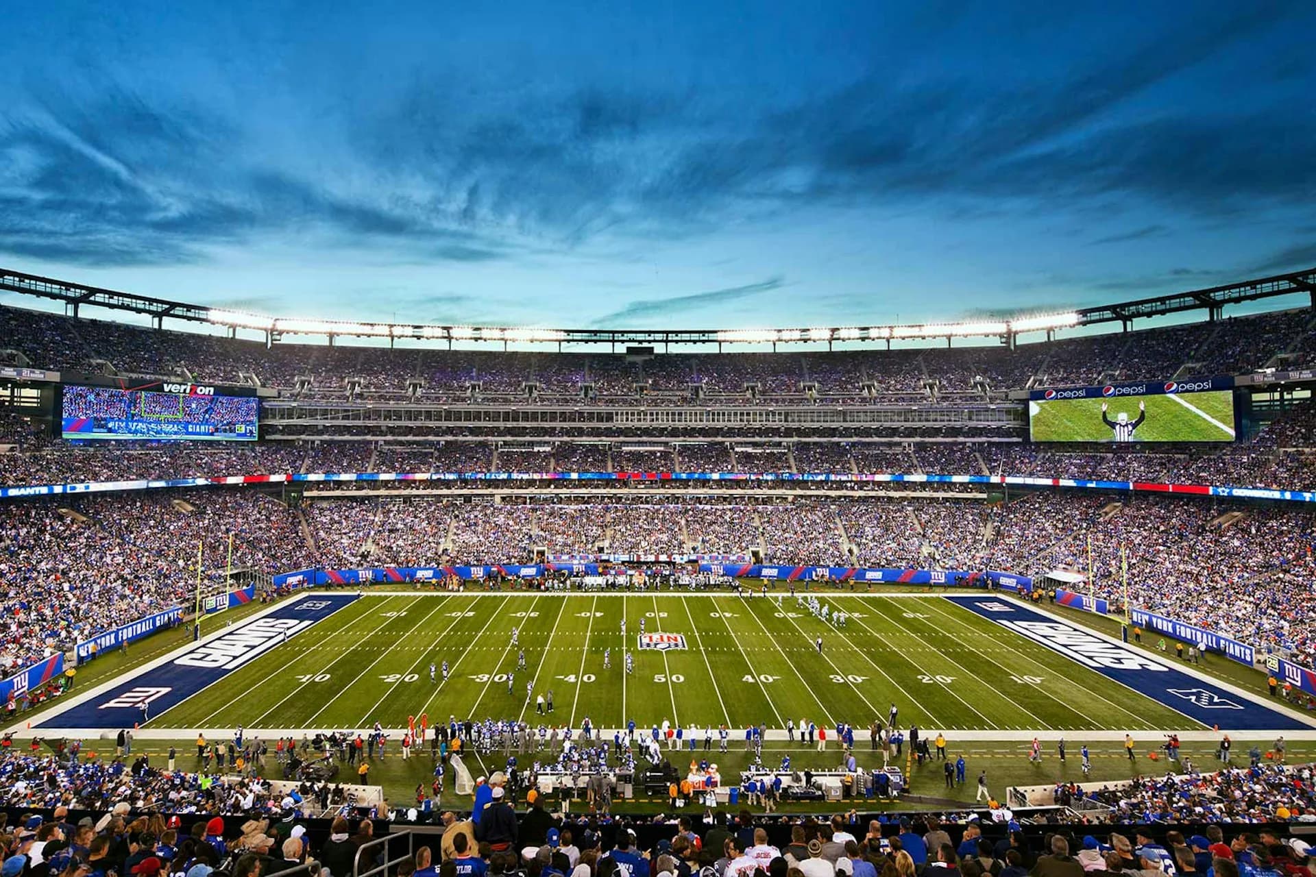 A packed stadium during a major sporting event with field markings, large video screens, and dramatic blue evening sky, showcasing architectural stadium design by Thornton Tomasetti.