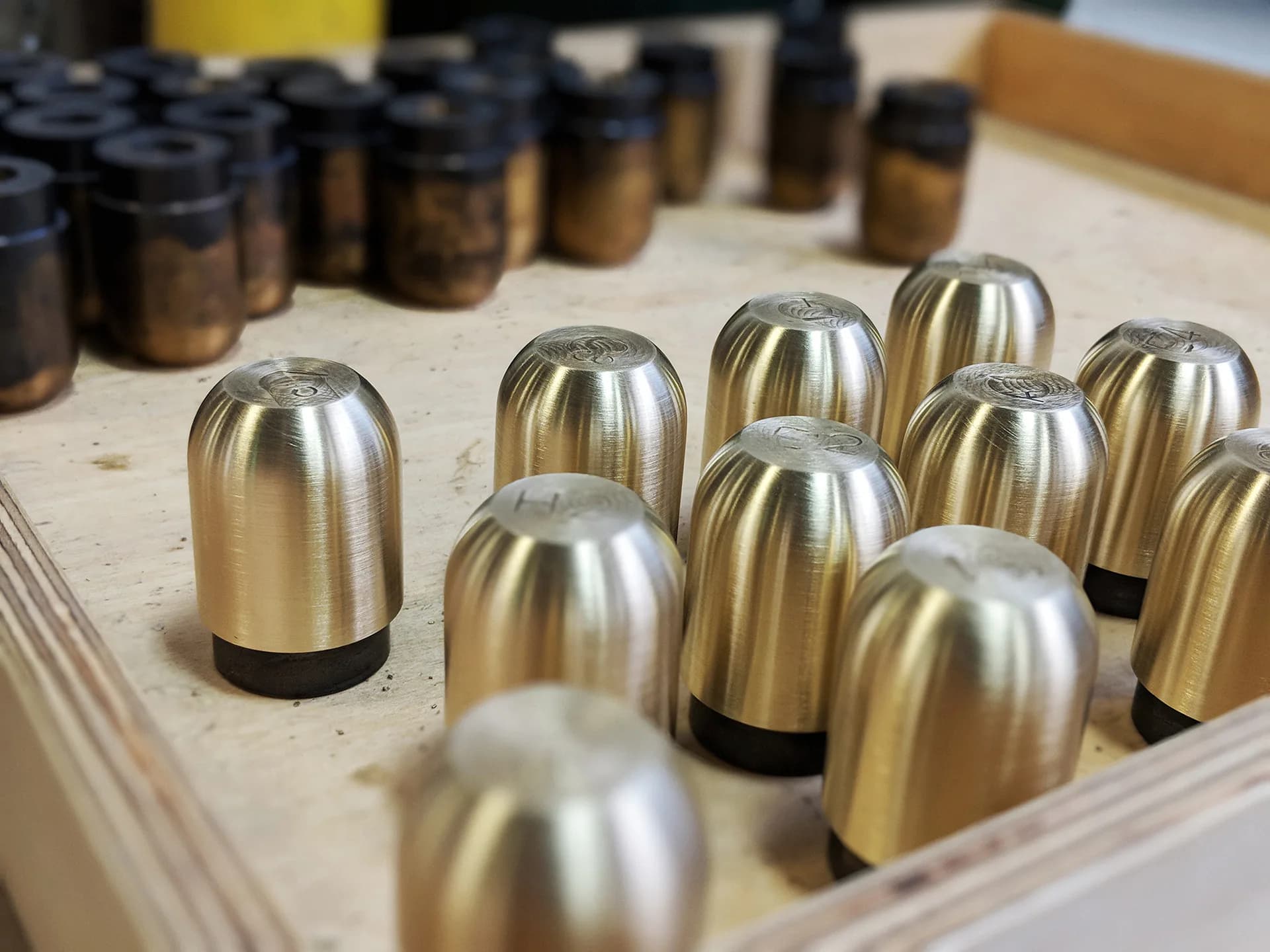 Polished brass bullet-shaped objects arranged in a wooden display tray with organized black containers in background, showcasing metalwork product design by Tietz-Baccon.