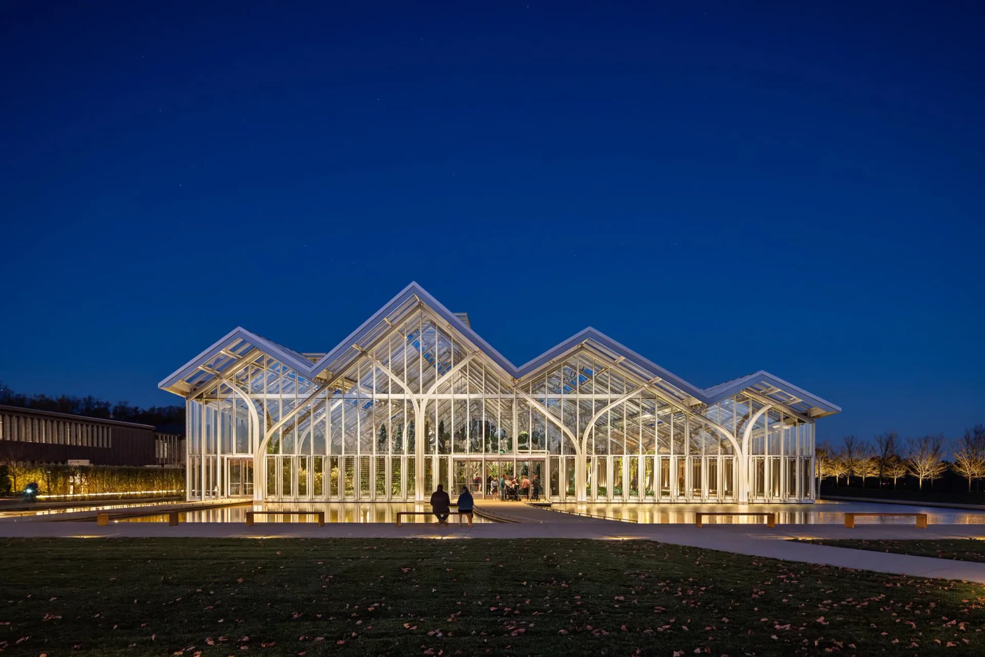 Illuminated modernist pavilion with white steel framework and peaked roof forms set against twilight sky, designed by Tillotson Design Associates.
