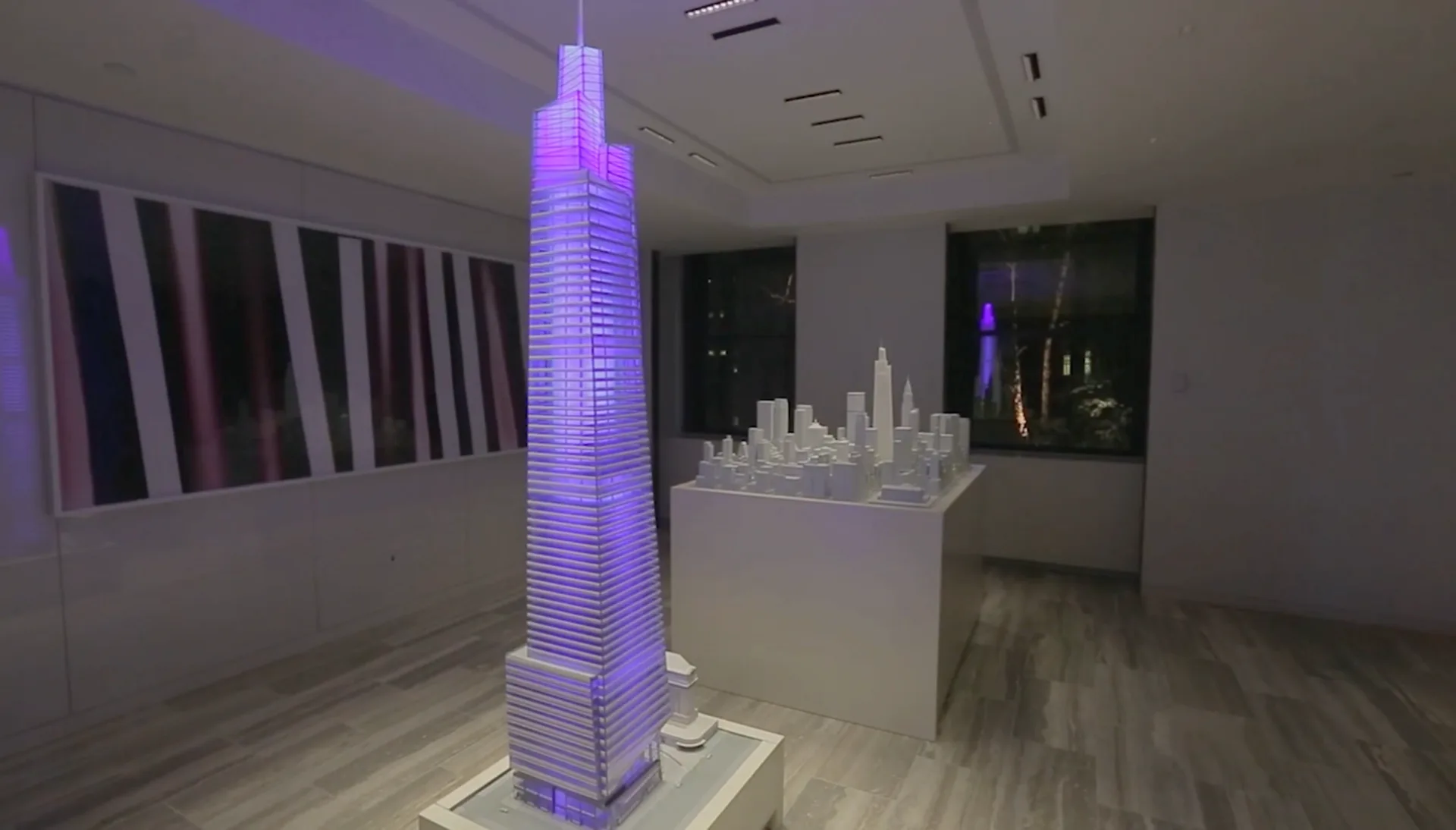 Modern gallery space with illuminated purple architectural model of a skyscraper on a pedestal, white cityscape relief on wall, and framed artwork, designed by Tirschwell & Co.