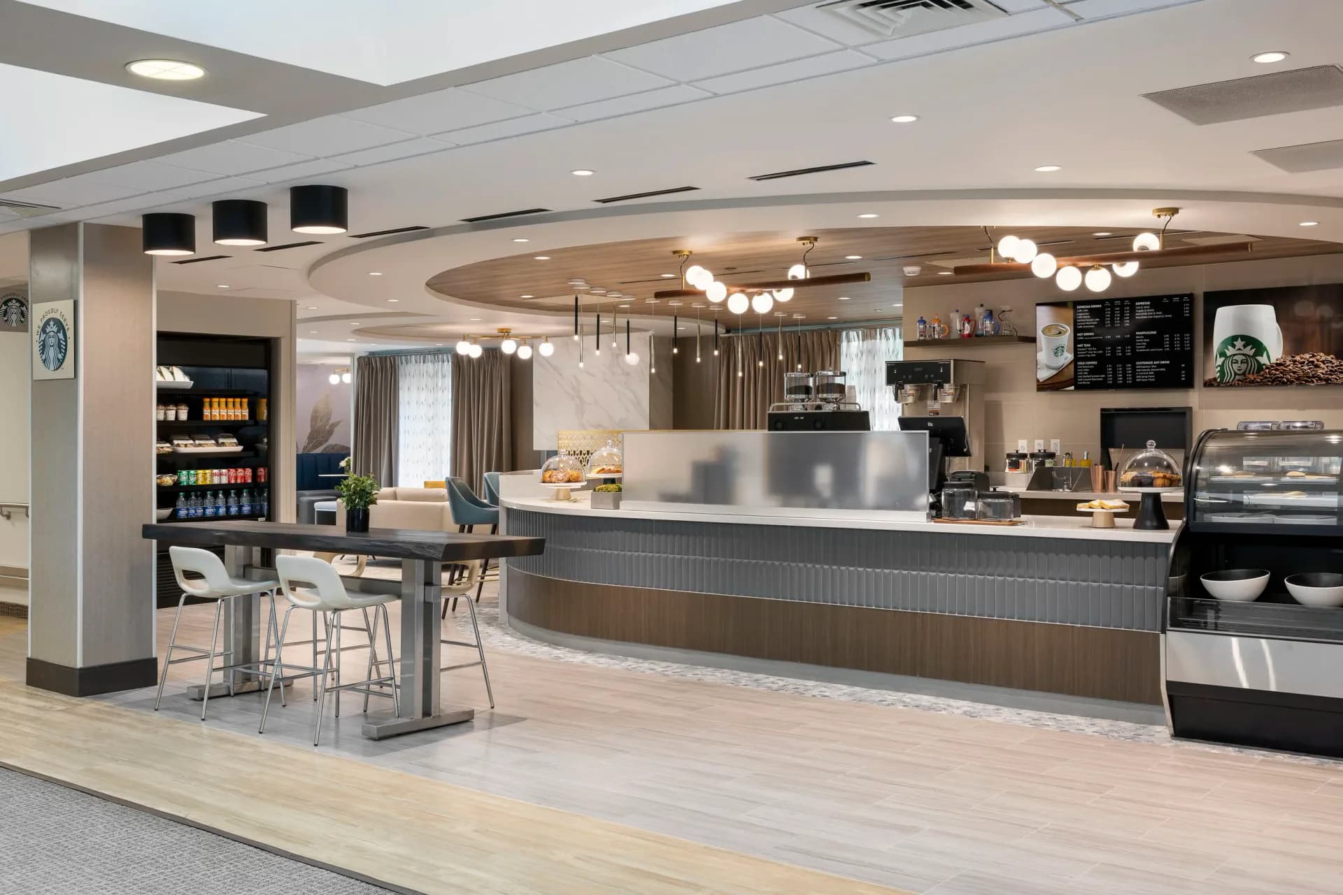 Modern café interior featuring a curved service counter with wood and dark finishes, pendant lighting, beverage cooler, and seating area with light wood flooring by TPD Architecture.