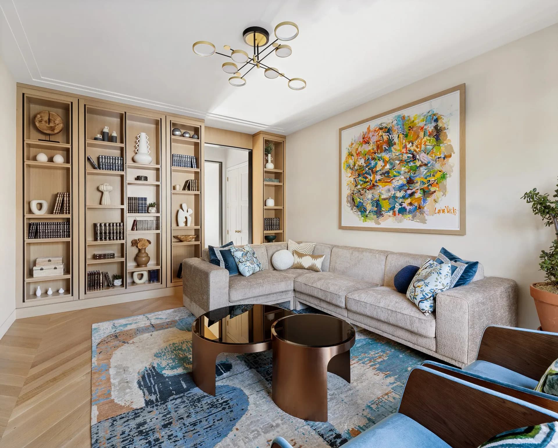 A light-filled living room features built-in wooden shelving, a gray sectional sofa with blue accents, a bronze cylindrical coffee table, and colorful artwork by Toby Kahn Interiors.