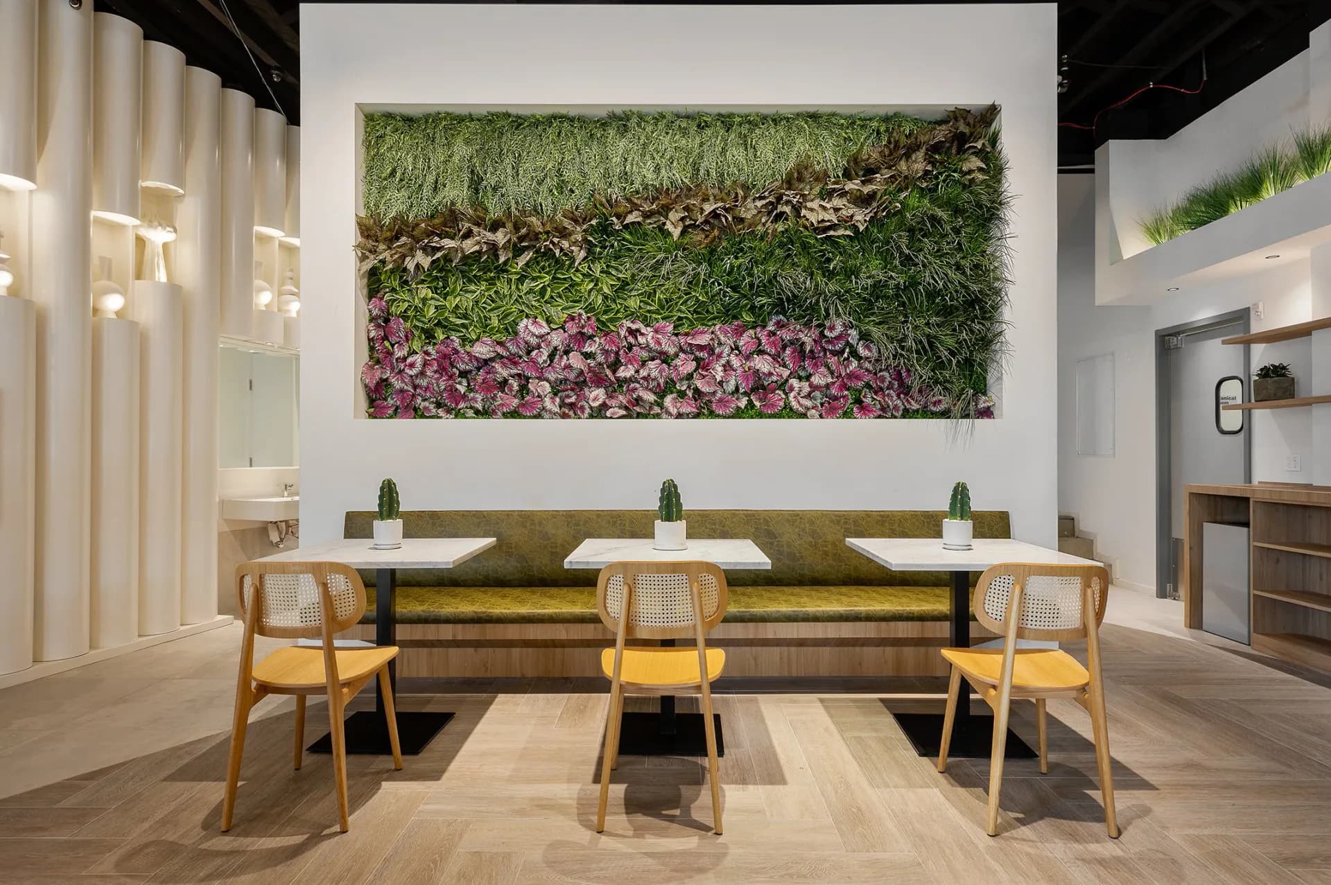 Modern dining space with olive-green banquette seating, wooden chairs, and a living green wall installation with purple flowers above, designed by Toby Kahn Interiors.