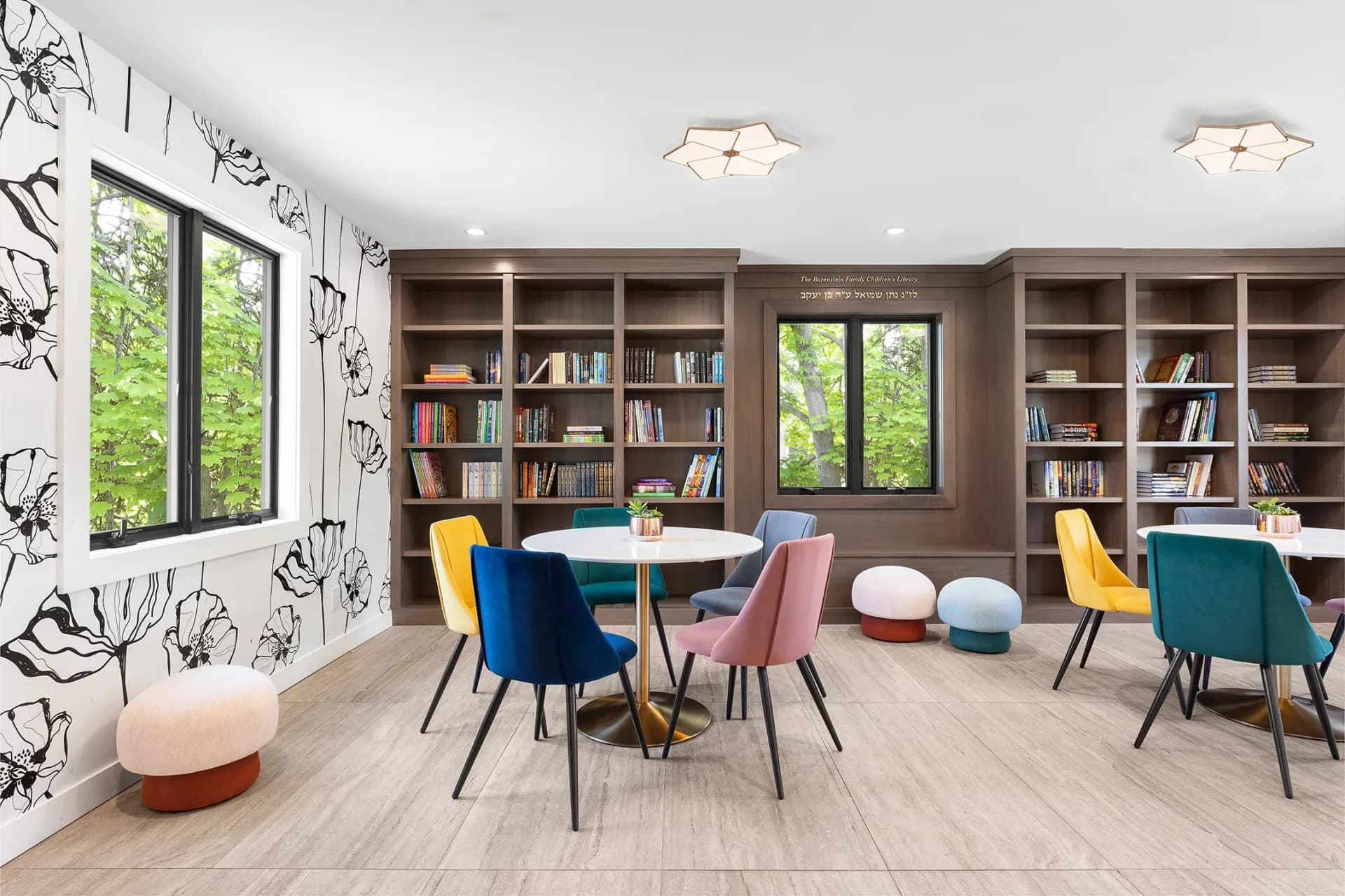 A bright library lounge with taupe built-in shelving, colorful mid-century chairs in navy, blush, and mustard, light wood flooring, and botanical-patterned accent wall by Toby Kahn Interiors.