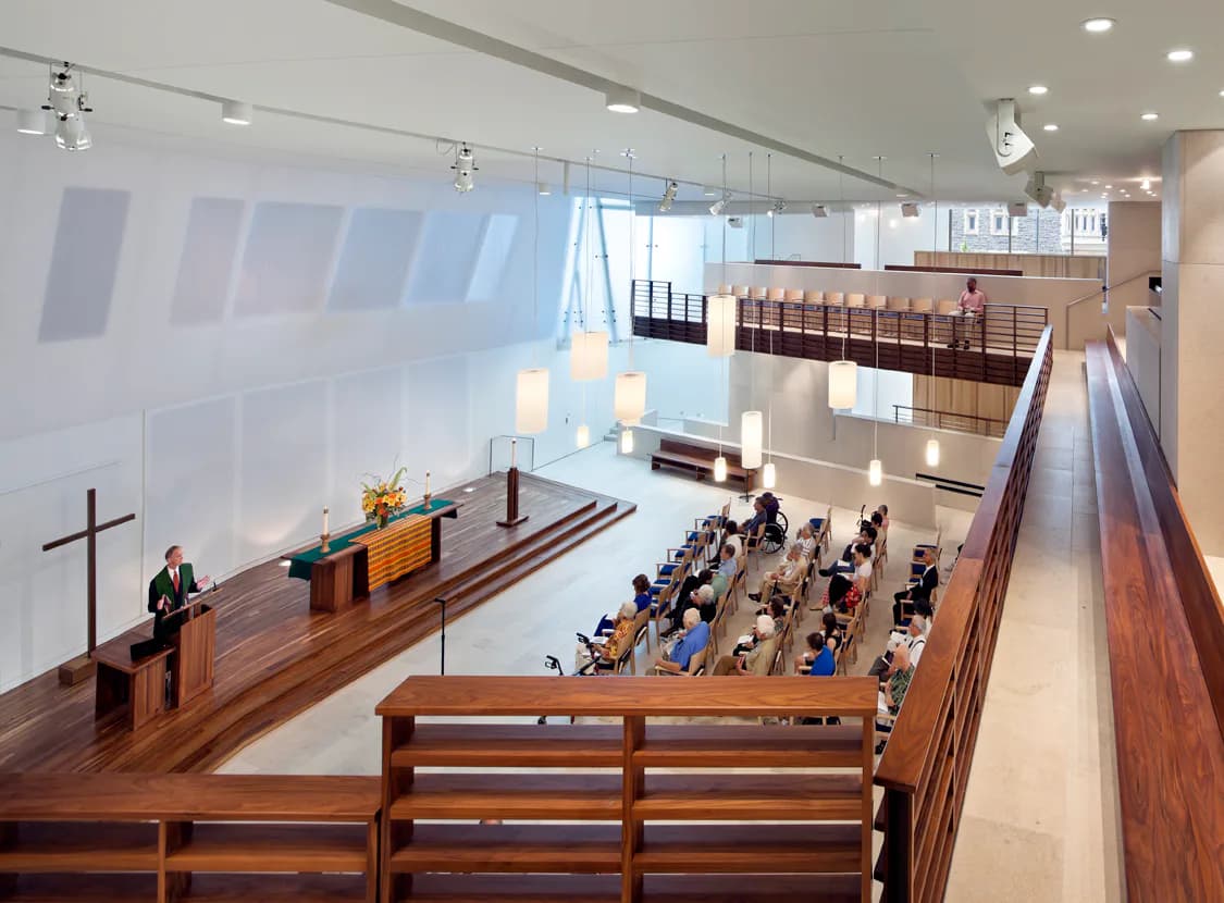Modern church interior with soaring white walls, wooden pews, and a raised altar platform with cross, designed by Tod Williams Billie Tsien Architects.