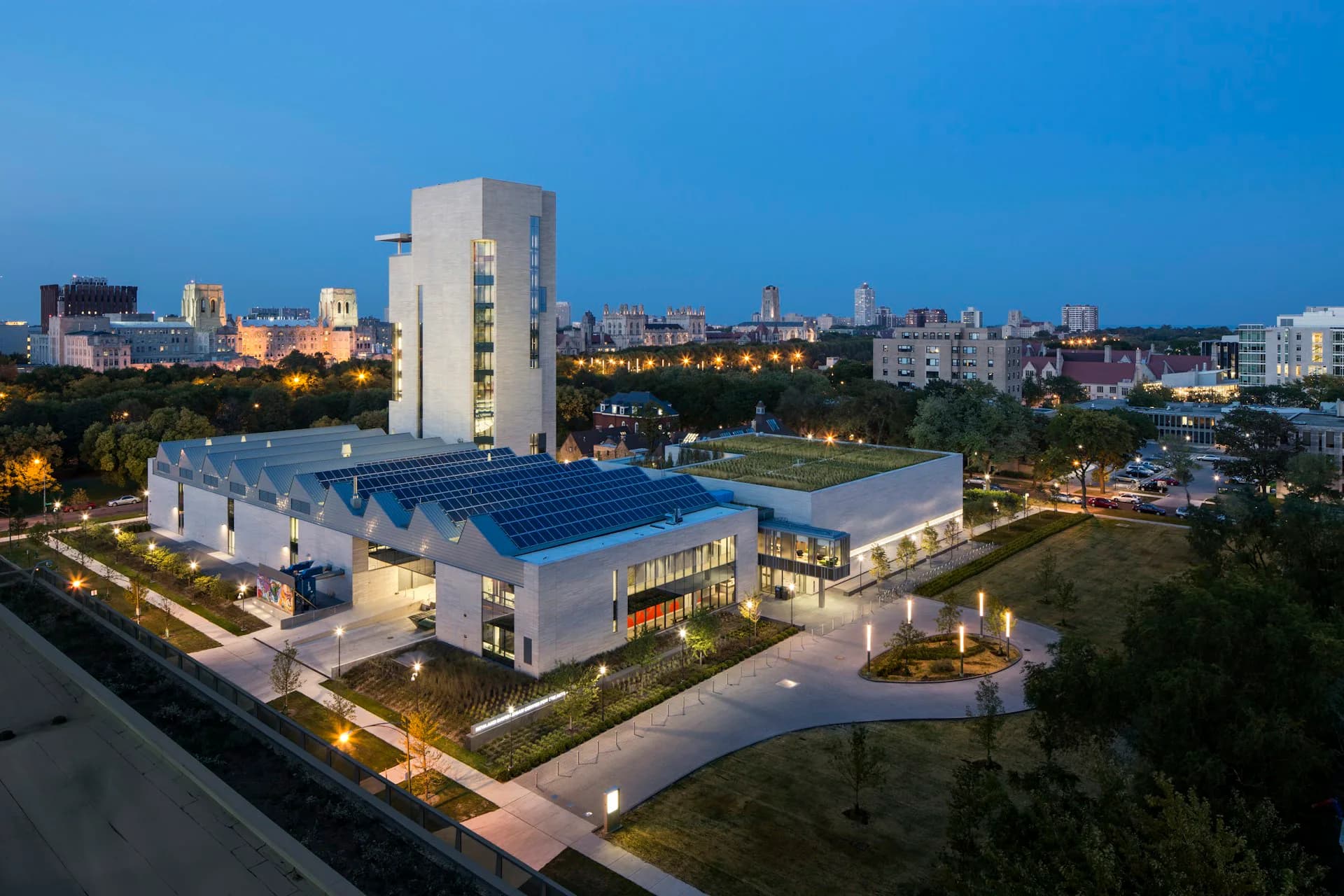 Modern institutional building with curved blue roof, illuminated white facades, and vertical tower element set against city skyline at dusk by Tod Williams Billie Tsien Architects | Partners.