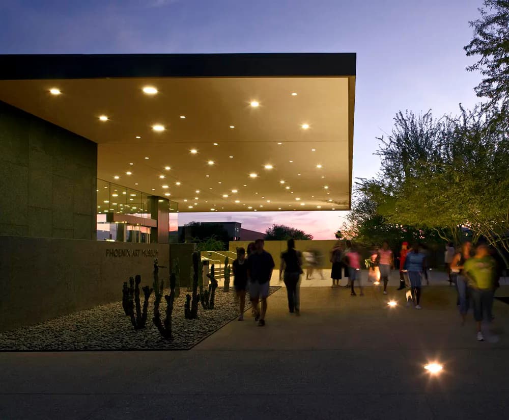 Evening exterior view of a modern pavilion with illuminated ceiling and warm lighting, surrounded by visitors on a plaza with integrated ground lights, designed by Tod Williams Billie Tsien Architects | Partners.