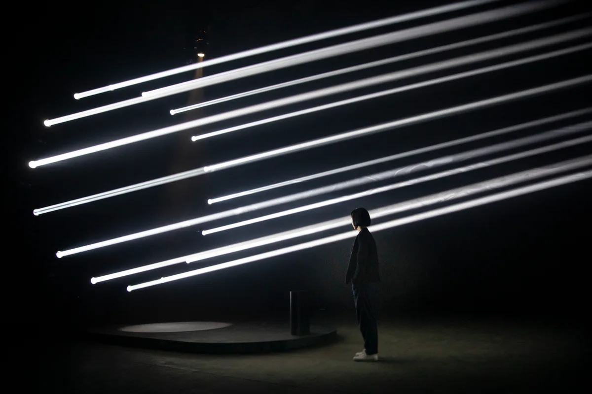Minimalist installation featuring a silhouetted figure standing beneath diagonal linear white neon lights against a dark background by Todd Bracher Studio.