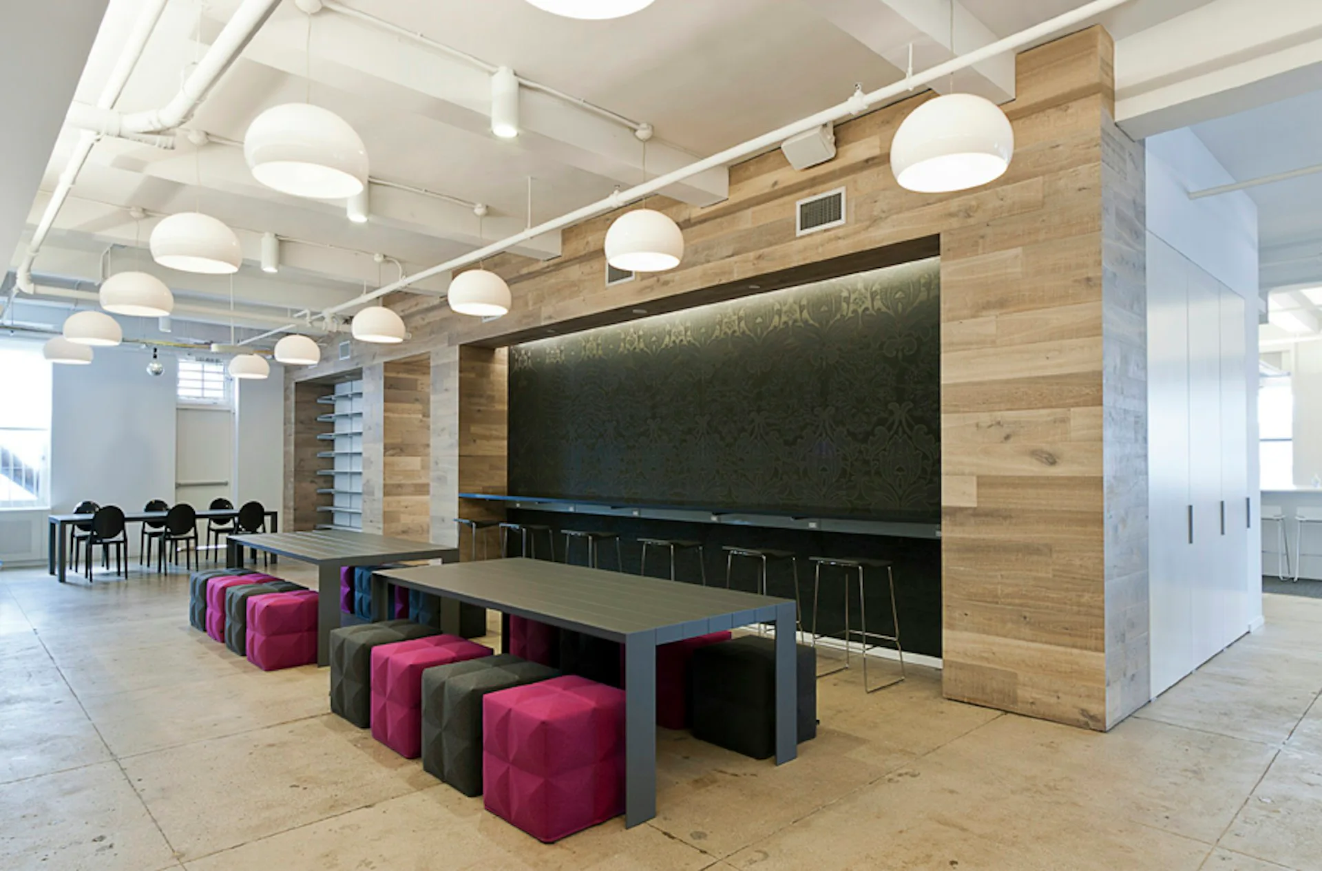 Modern office breakroom with large blackboard wall, wooden accent paneling, colorful modular seating, and contemporary lighting by TPG Architecture LLP.