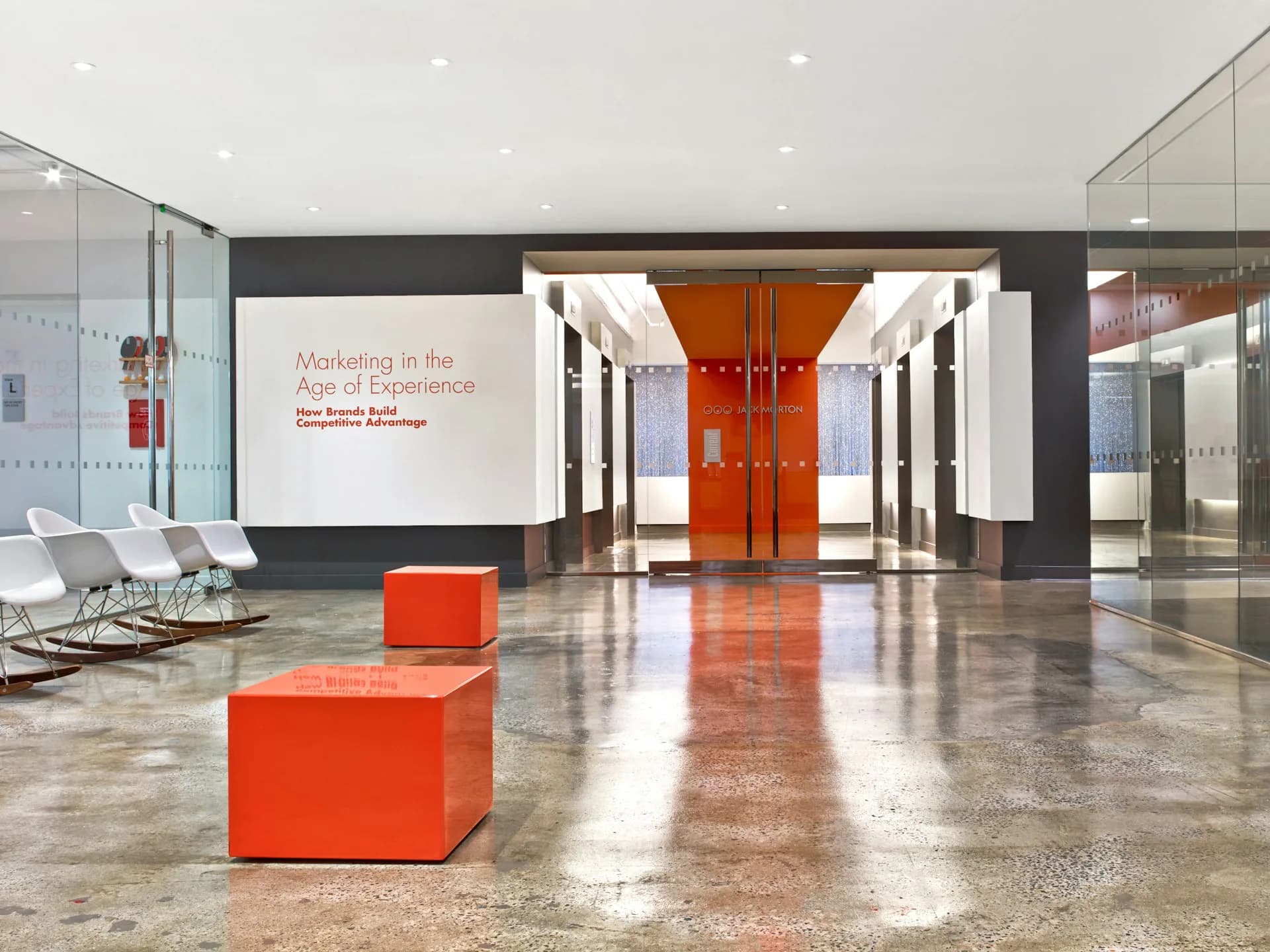 Modern lobby with polished concrete floors, red cubic seating elements, orange doors, and dark accent walls by TPG Architecture LLP.