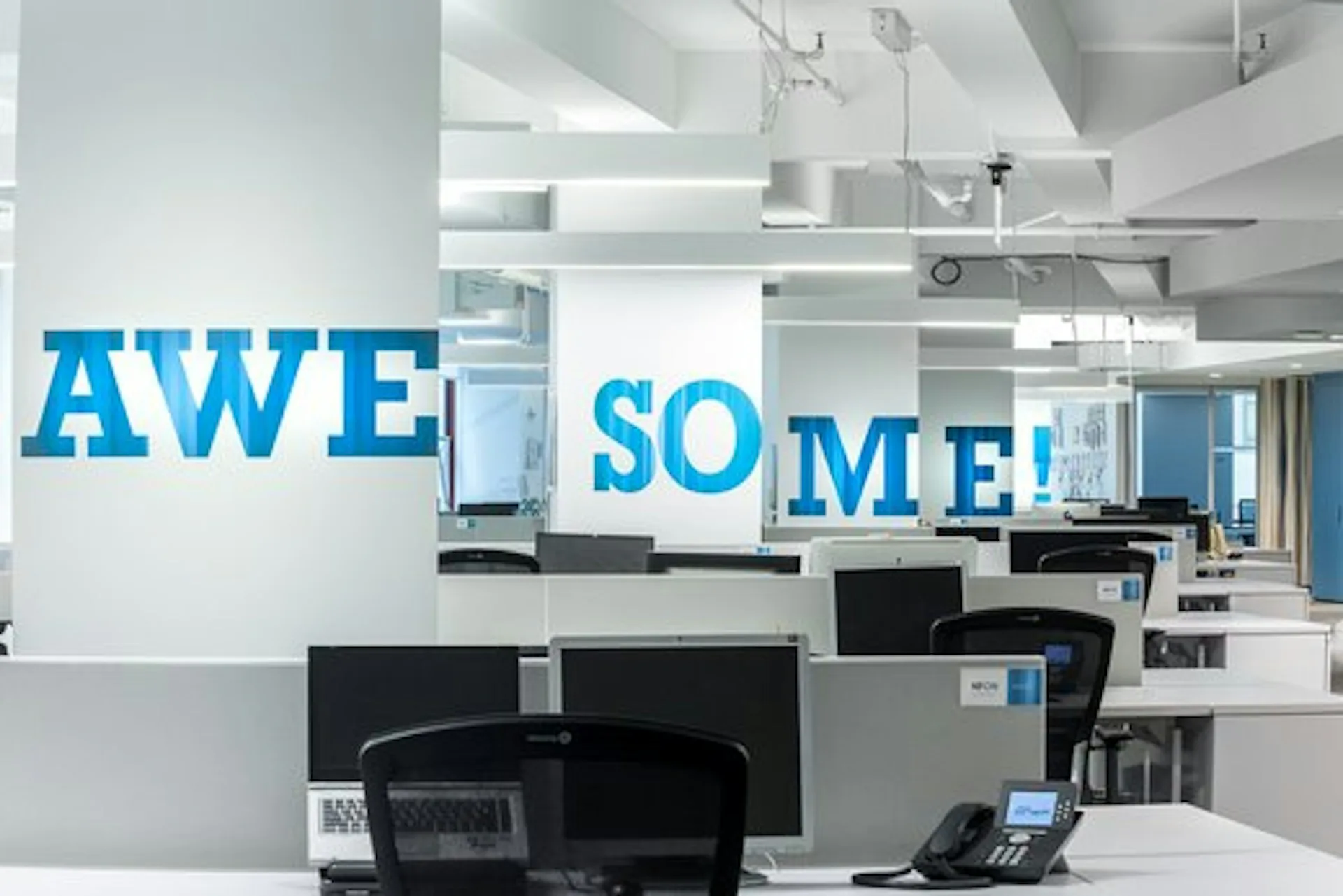 Modern office space with white ceilings, dark computer workstations, and blue 'AWESOME!' wall typography by TPG Architecture LLP.
