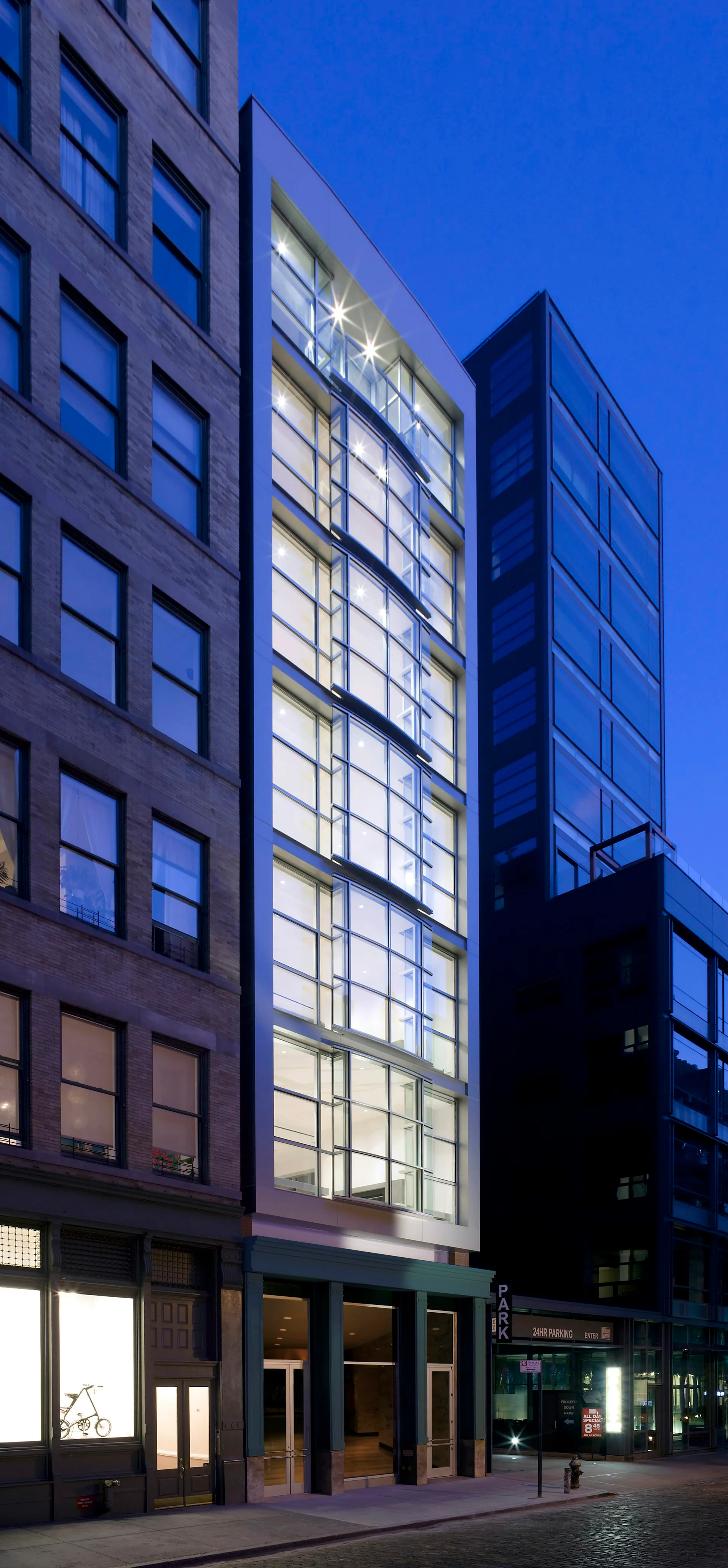 Illuminated modern mixed-use building facade at dusk with gridded glass windows, horizontal brass accents, and dark stone cladding by TRA studio Architecture PLLC, New York City.
