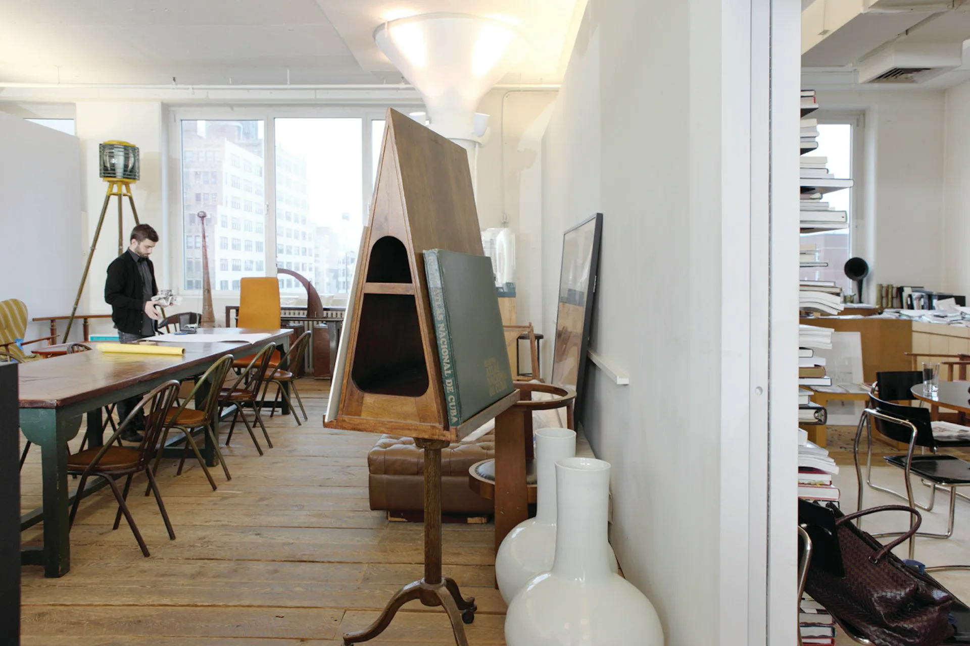Modern office space with wooden flooring, white walls, and large windows; features a work table with chairs, sculptural wooden elements, and glass partitions creating an open-plan layout by TsAO & McKOWN Architects.