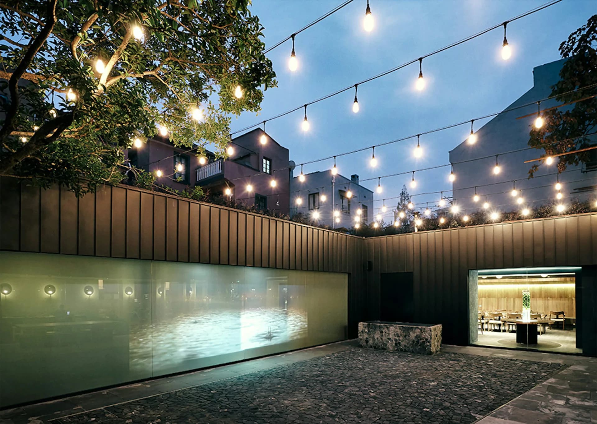 Modern courtyard with frosted glass walls, string lighting overhead, and contemporary buildings beyond, designed by TsAO & McKOWN Architects.