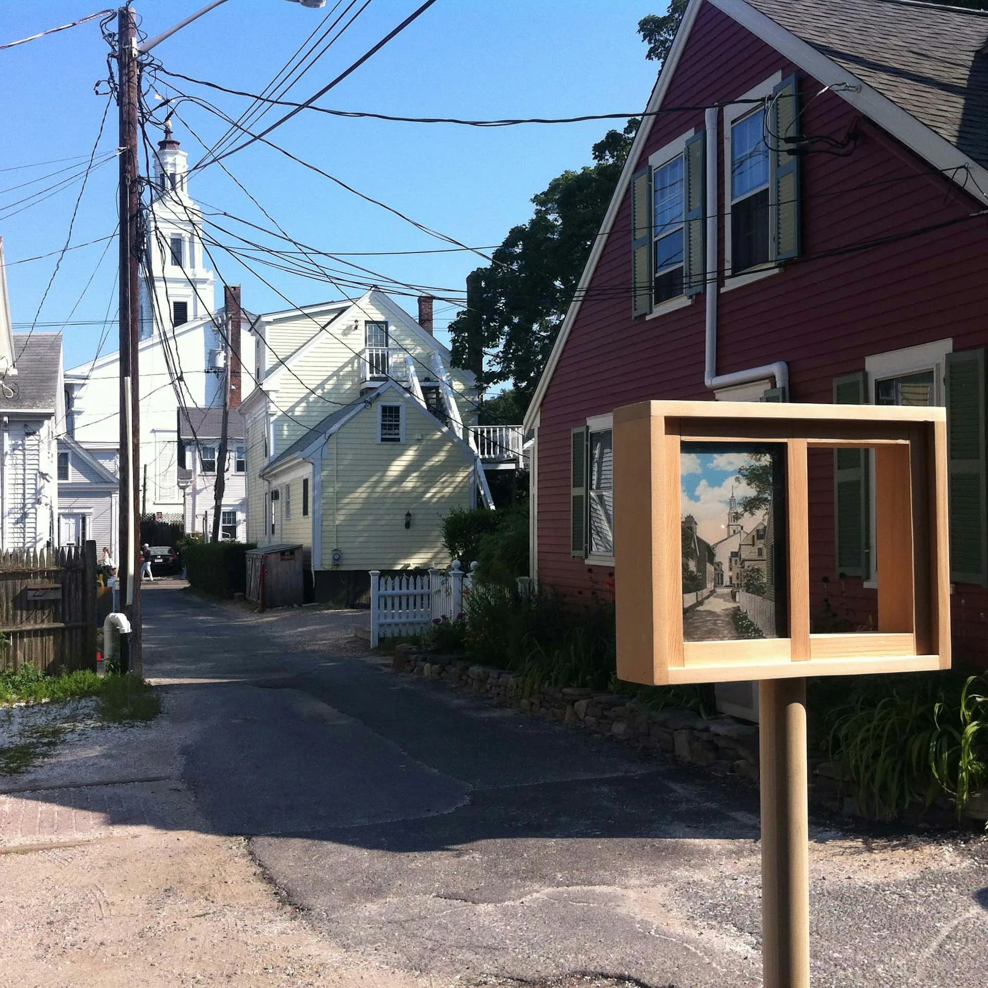 A wooden library box mounted on a residential street with an orange frame and glass doors, displaying books, situated among New England colonial-style homes by TsAO & McKOWN Architects.