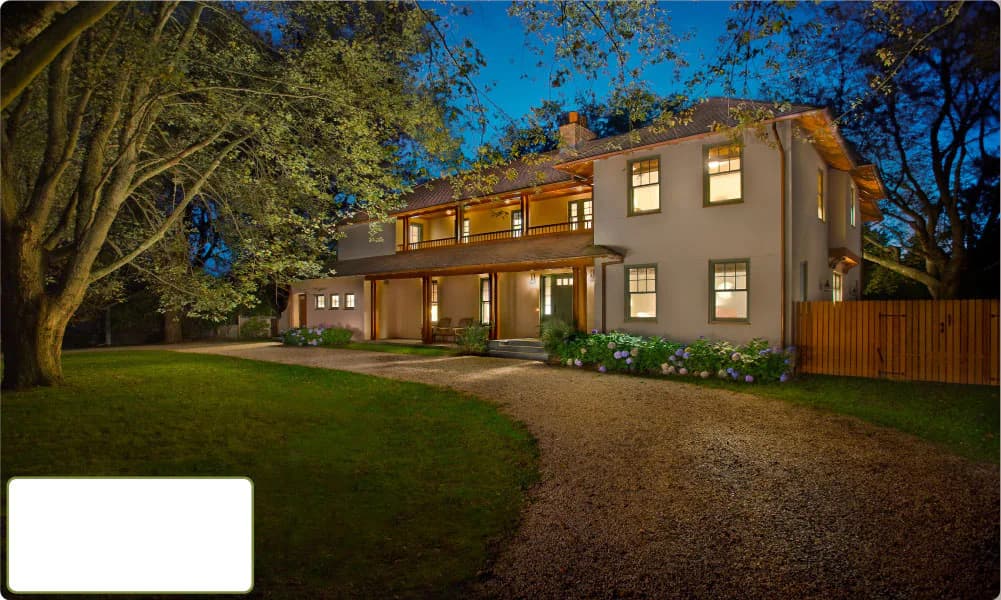 Contemporary two-story residential home with warm taupe exterior, illuminated windows at dusk, manicured lawn, gravel driveway, and mature trees by Turino Kalberer Architects.