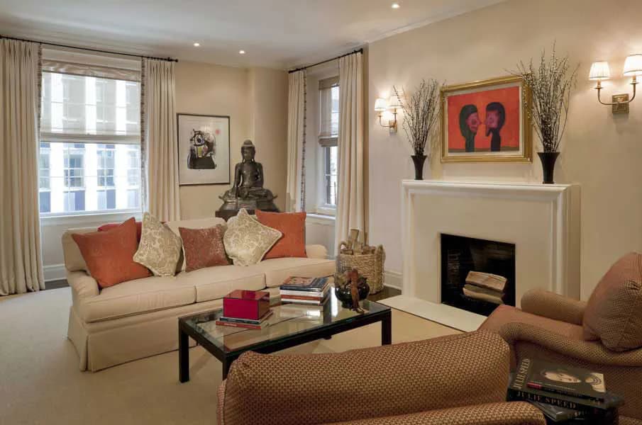 Elegant living room with cream sofa, orange accent pillows, fireplace with red artwork, patterned armchair, and large windows with draped curtains by Turino Kalberer Architects.