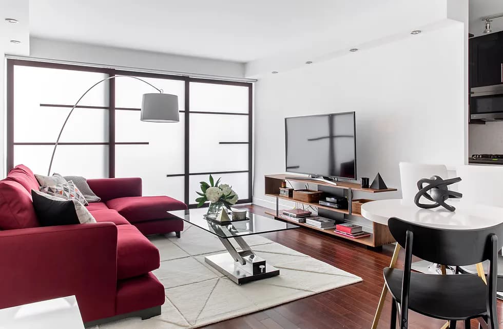 Modern living room with burgundy sectional sofa, wooden floor, white walls, and large gridded windows overlooking a city, designed by Twelfth Street Interiors.