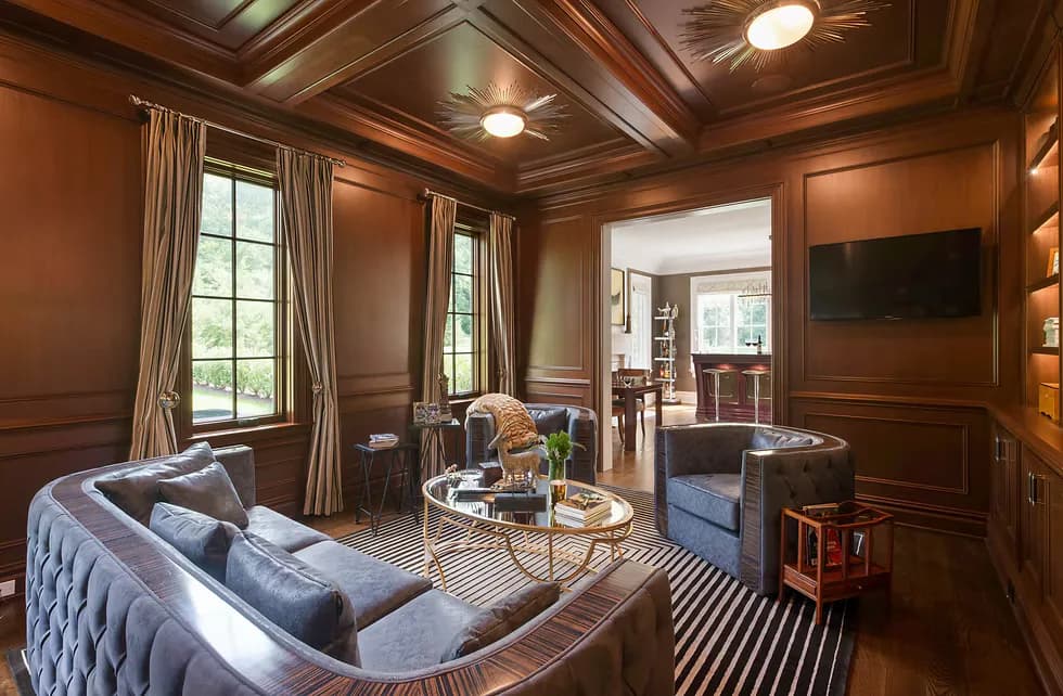 Luxurious wood-paneled living room with blue upholstered sofa, striped area rug, coffered ceiling, and large windows overlooking greenery by Twelfth Street Interiors.