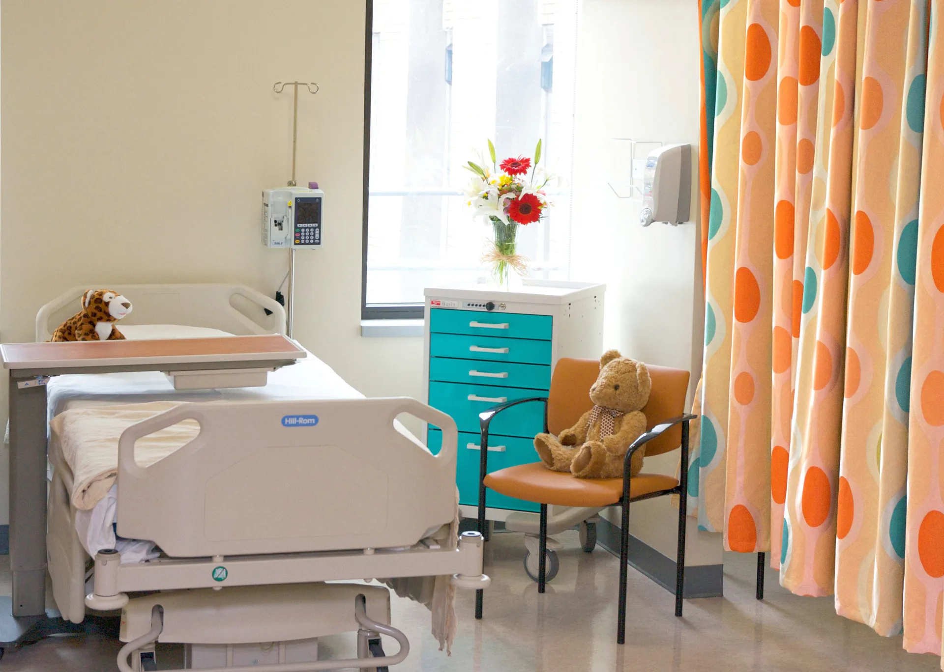 Pediatric hospital room with turquoise medical cabinetry, colorful patterned curtains, soft furnishings, and welcoming décor by Twine Architectural Studio.