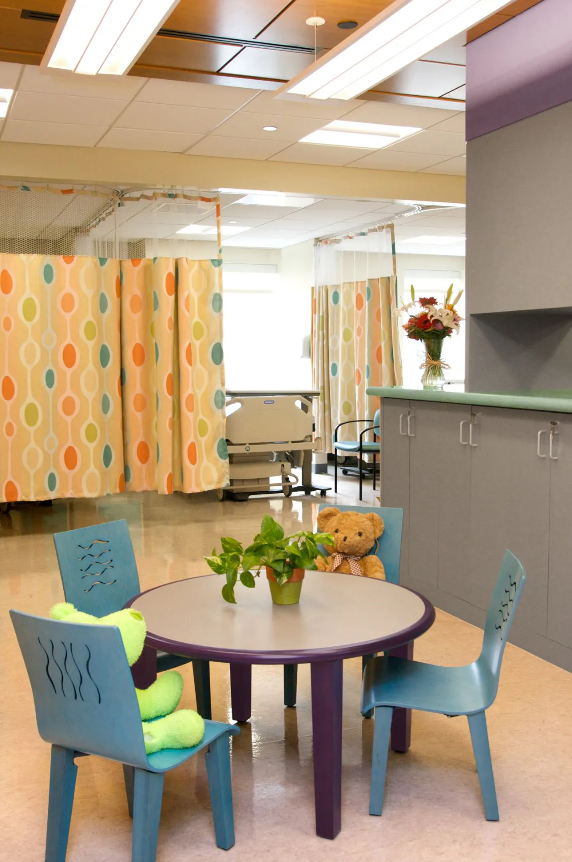 Pediatric clinic interior with colorful patterned privacy curtains, purple round table with blue chairs, potted plant, and teddy bear by Twine Architectural Studio.