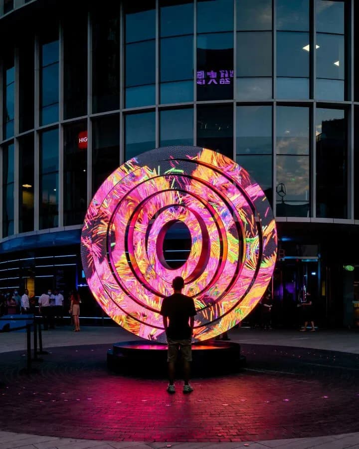 Large illuminated circular neon installation with concentric rings in pink, magenta, and gold tones, displayed in an urban plaza at dusk by UAP (Urban Art Projects).
