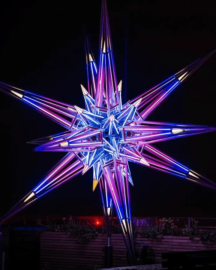 Illuminated neon star sculpture with radiating spikes in blue, purple, and gold tones against a dark night sky by UAP (Urban Art Projects).