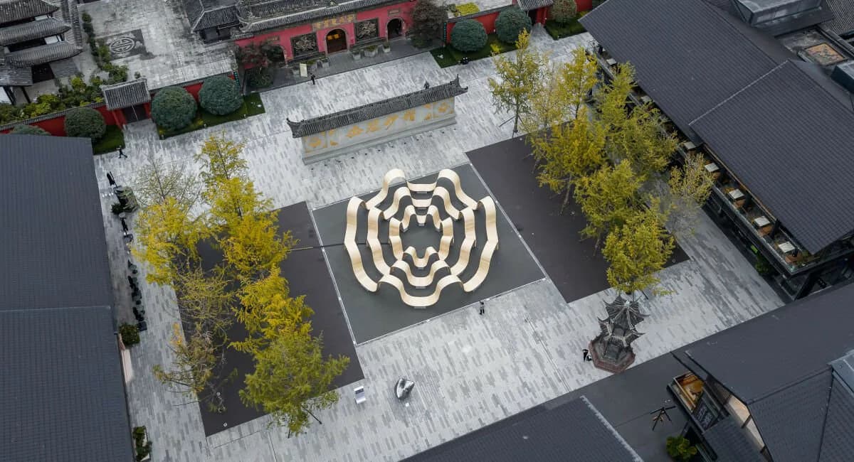 Aerial view of a minimalist plaza courtyard featuring a geometric concentric circular pattern in white inlay on dark pavement, surrounded by mature trees and modern architecture by UAP (Urban Art Projects).