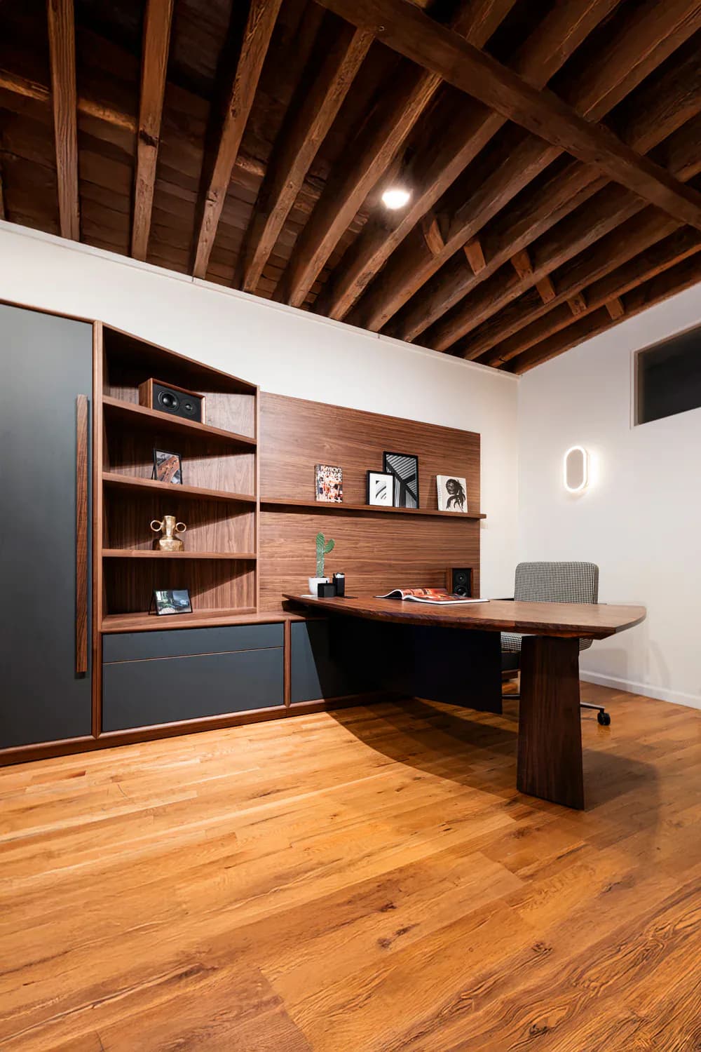 Modern home office featuring walnut desk and shelving with black accents, exposed wood beam ceiling, and warm wood flooring by Uhuru Design.