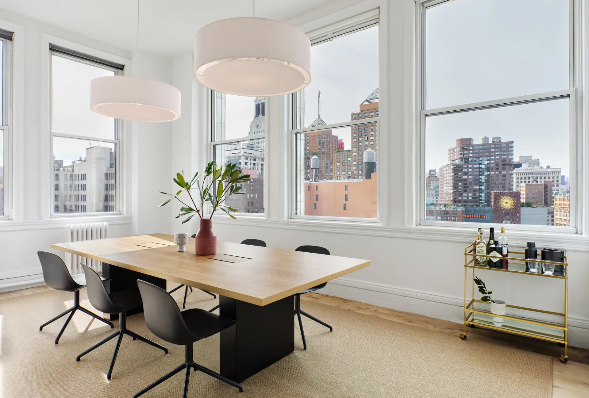 Modern dining space with natural wood table, black chairs, and cylindrical pendant lights overlooking a city skyline by Uhuru Design.