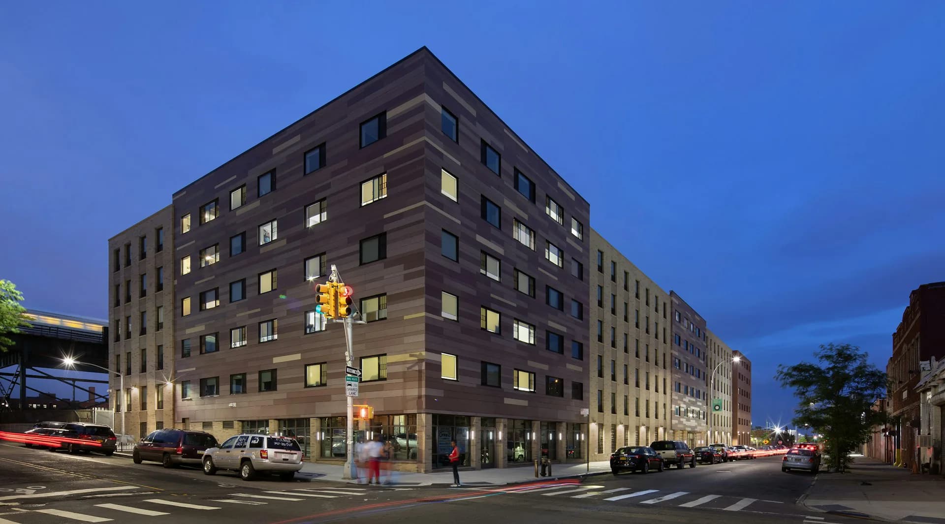 Modern residential building with dark brick facade and illuminated windows at dusk, corner intersection with street-level retail by Urban Architectural Initiatives.