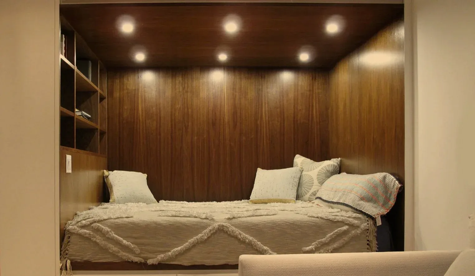 Recessed bedroom nook with wood-paneled walls, built-in bed with cream bedding, floating shelves, and warm ambient lighting by Urban Homecraft.