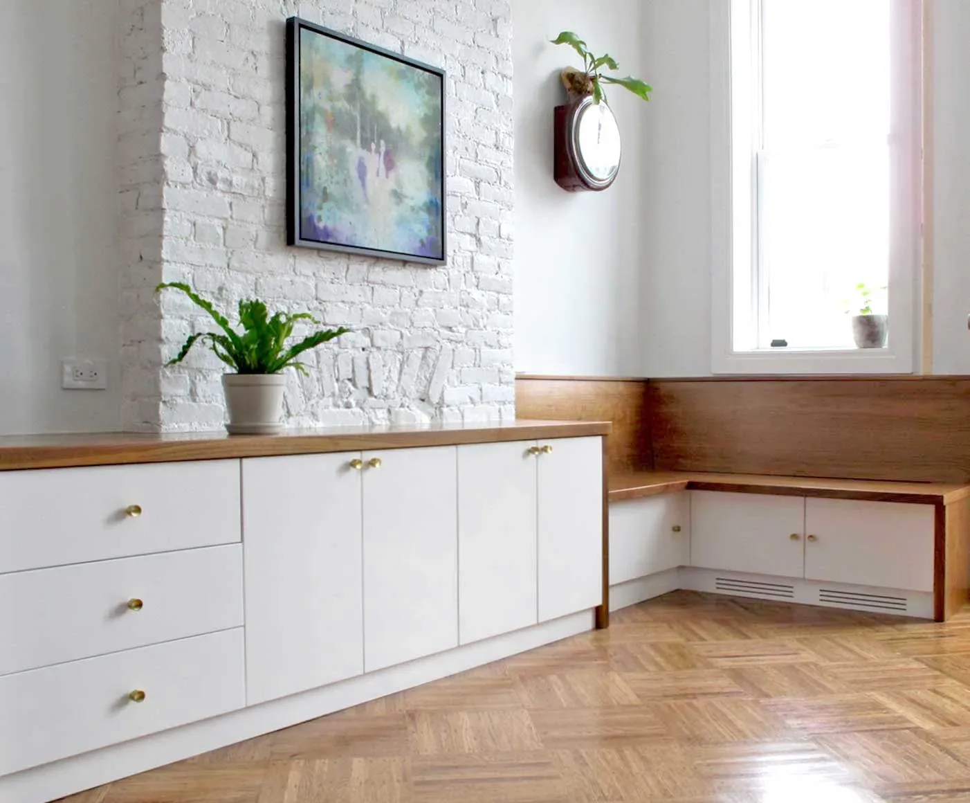Modern residential interior with white painted brick wall, wood-trimmed cabinetry, herringbone flooring, potted plants, and framed artwork by Urban Homecraft.