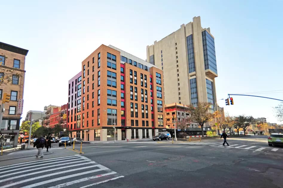 Urban street intersection with modern brick residential buildings in warm orange and red tones, featuring ground-floor retail and mid-rise architecture by Urban Quotient, PC.