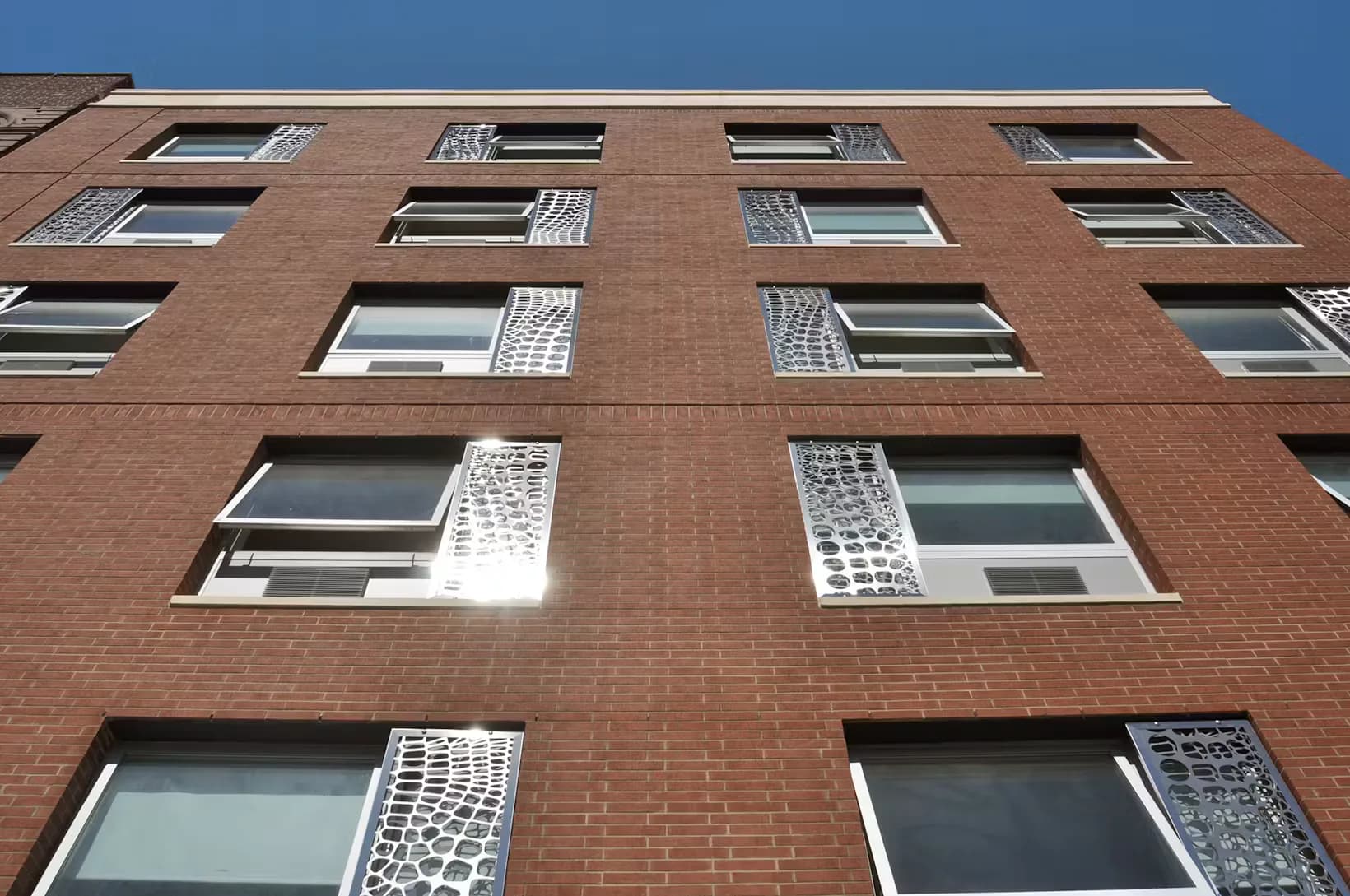 Brick residential building facade with regularly spaced windows and white decorative metal grilles, photographed from street level by Urban Quotient, PC.