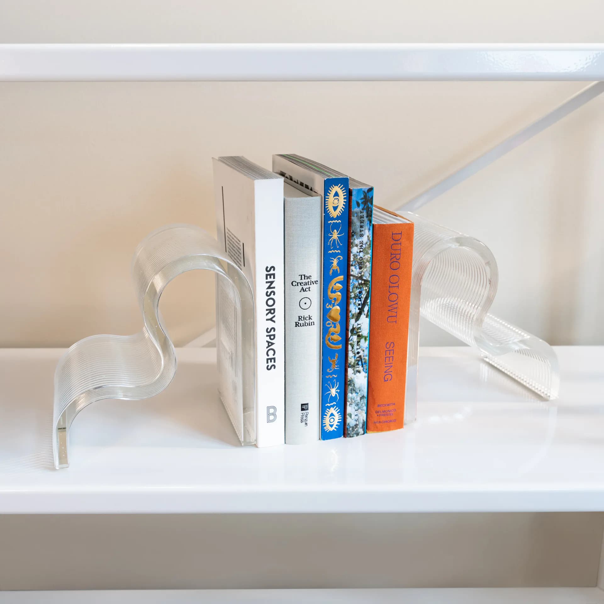 Minimalist white shelving display with clear acrylic bookends holding a curated stack of colorful books, featuring clean lines and modern styling by UrbanGlass.