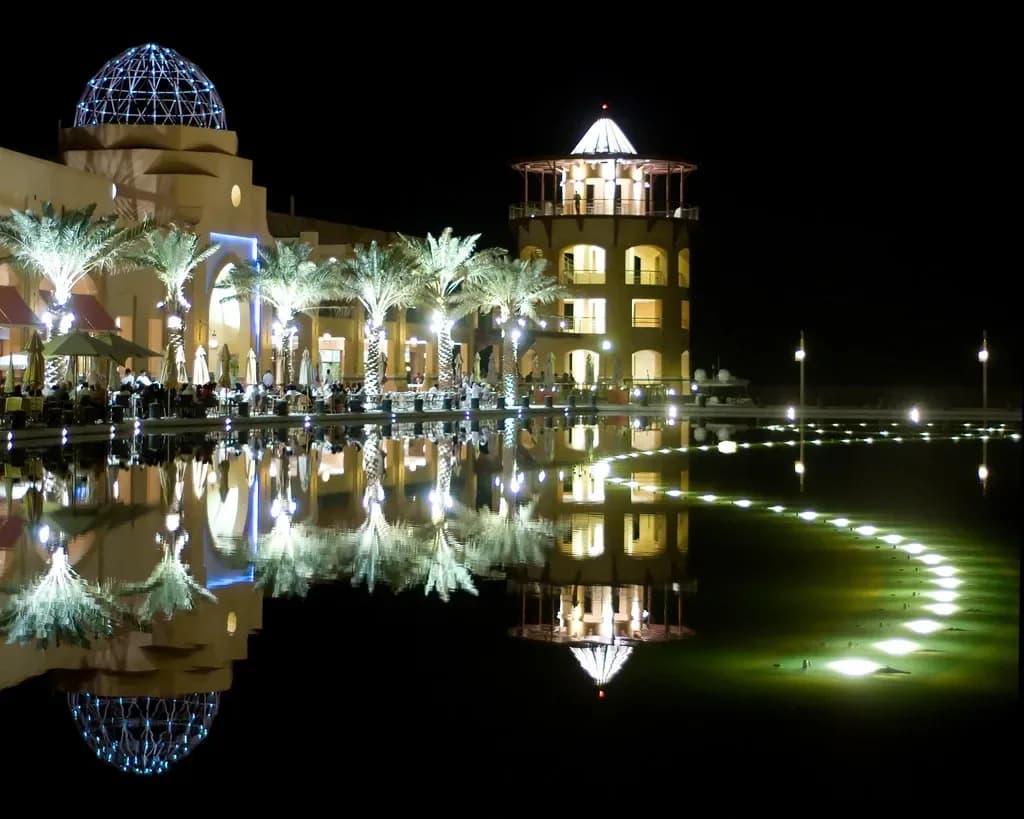 Nighttime architectural lighting design of a waterfront plaza with illuminated domed and tower structures reflected in still water, featuring palm trees and pathway lighting by US Lighting Consultants.