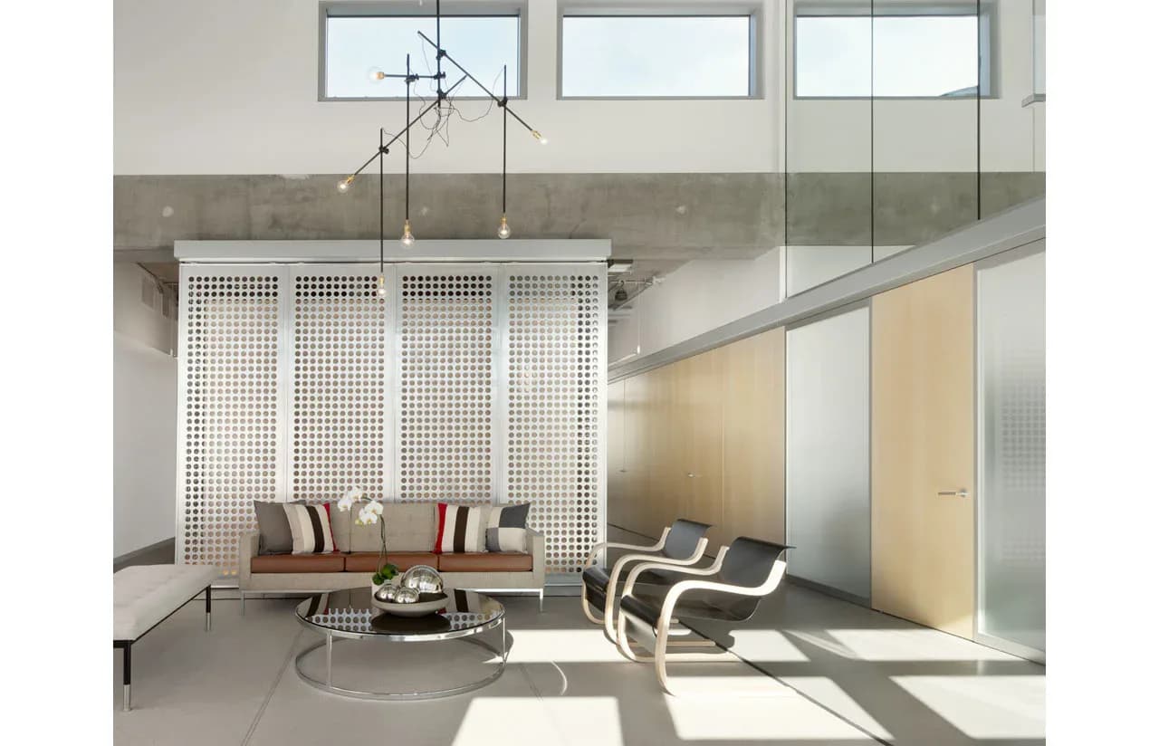 Modern loft interior with concrete walls, geometric pendant lighting, minimalist furniture, and floor-to-ceiling windows flooding the space with natural light, designed by Ventresca Lighting Designers.
