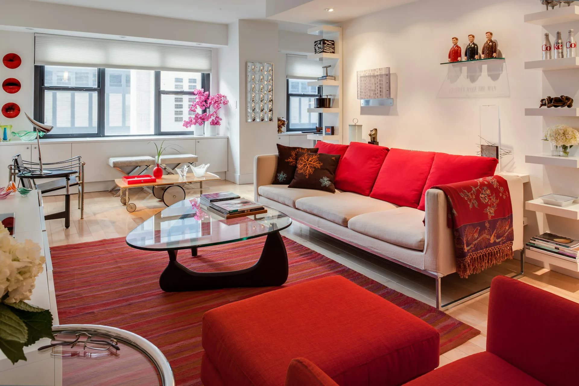 Modern living room with gray sofa, red pillows and ottoman, red area rug, black coffee table, and large windows with city views, designed by Victoria Benatar ARCHITECT PLLC.