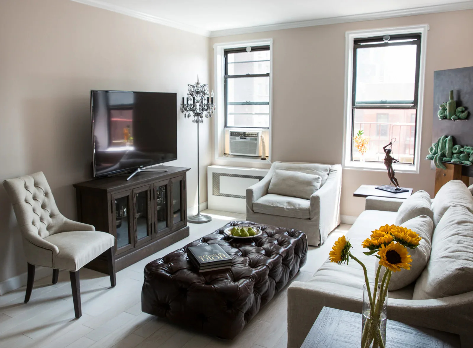 Contemporary living room with neutral palette, dark media console, tufted black ottoman, cream upholstered chair, and natural light from large windows, designed by Victoria Benatar ARCHITECT PLLC.