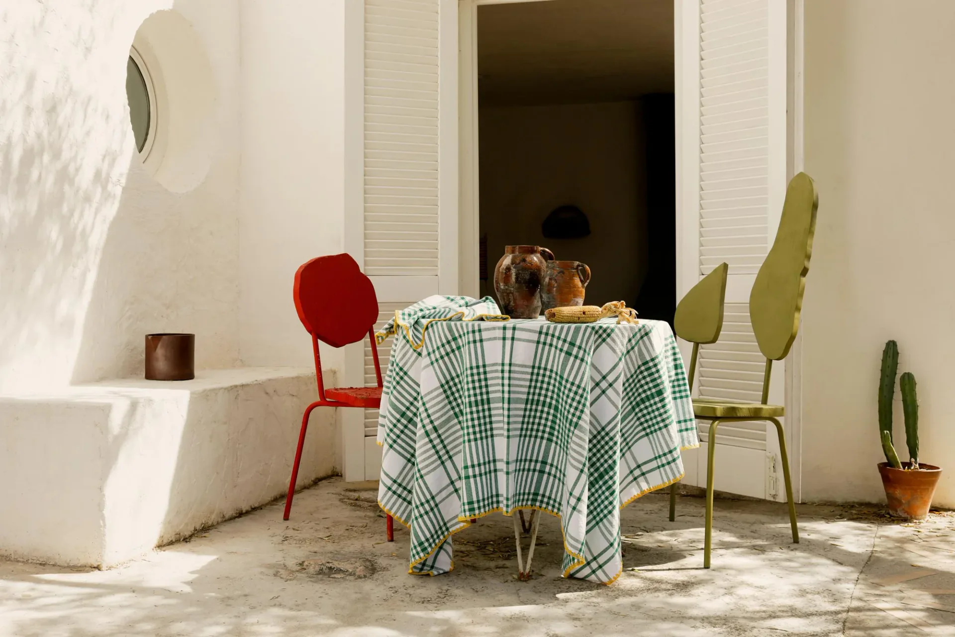 Bright Mediterranean courtyard with white walls, checkered tablecloth-draped table, red and olive chairs, potted cacti, and louvered doors by Viso Project.