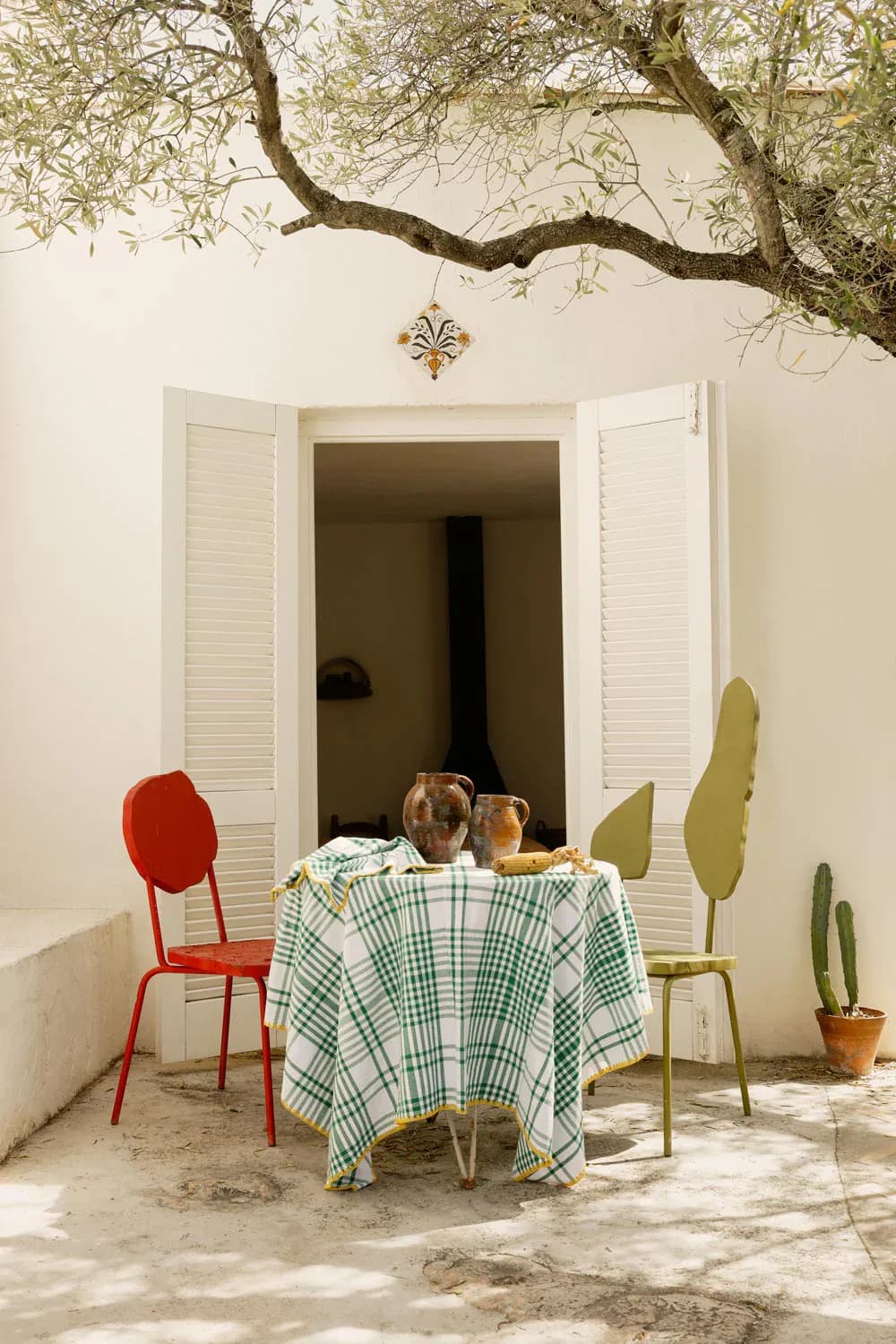 Minimalist courtyard with white walls, draped checkered table, red and olive chairs, potted cacti, and tree overhead by Viso Project.